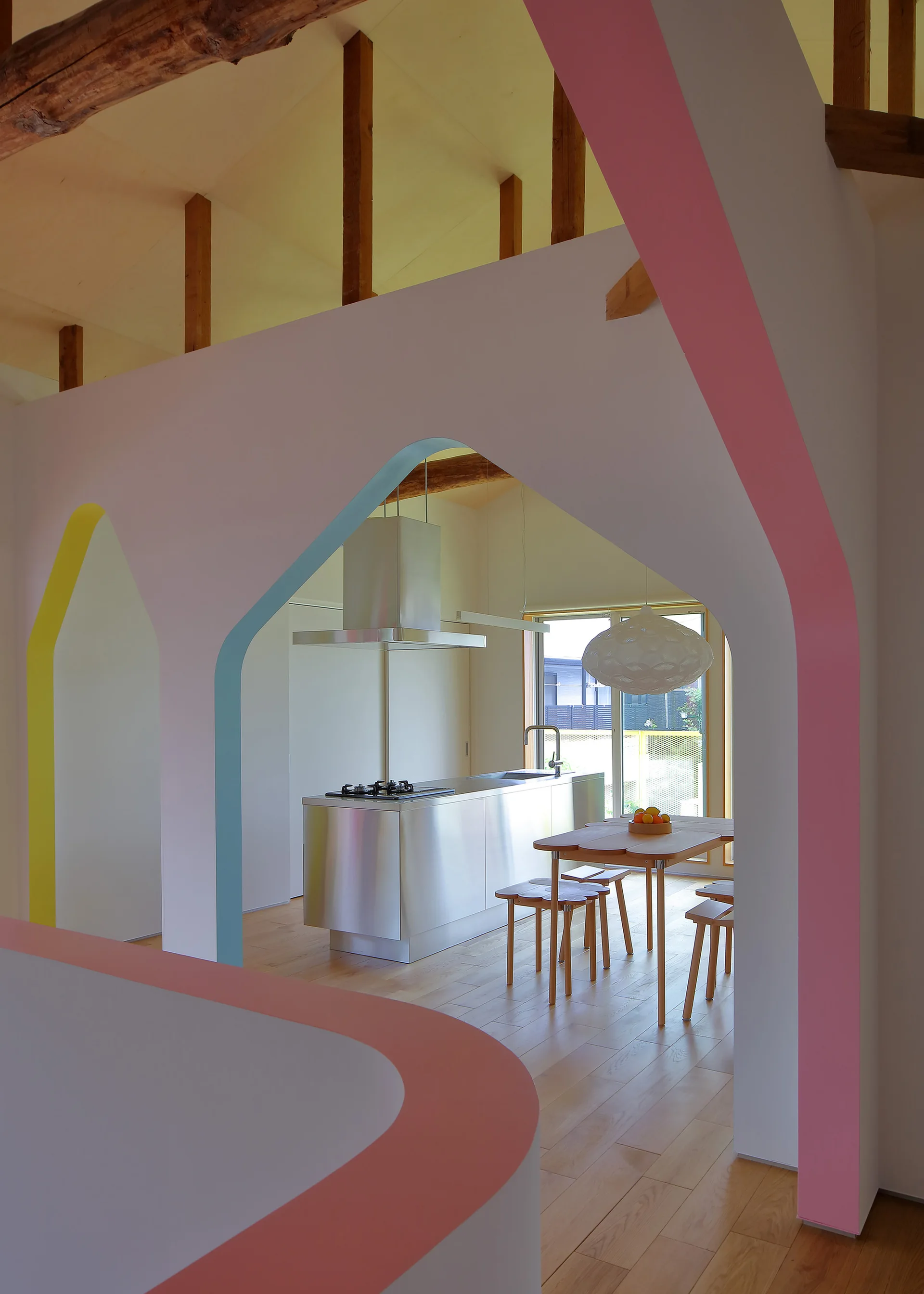 A stainless steel kitchen island and wooden dining set are framed by multiple white structural archways detailed with pink, blue, and yellow painted trim.