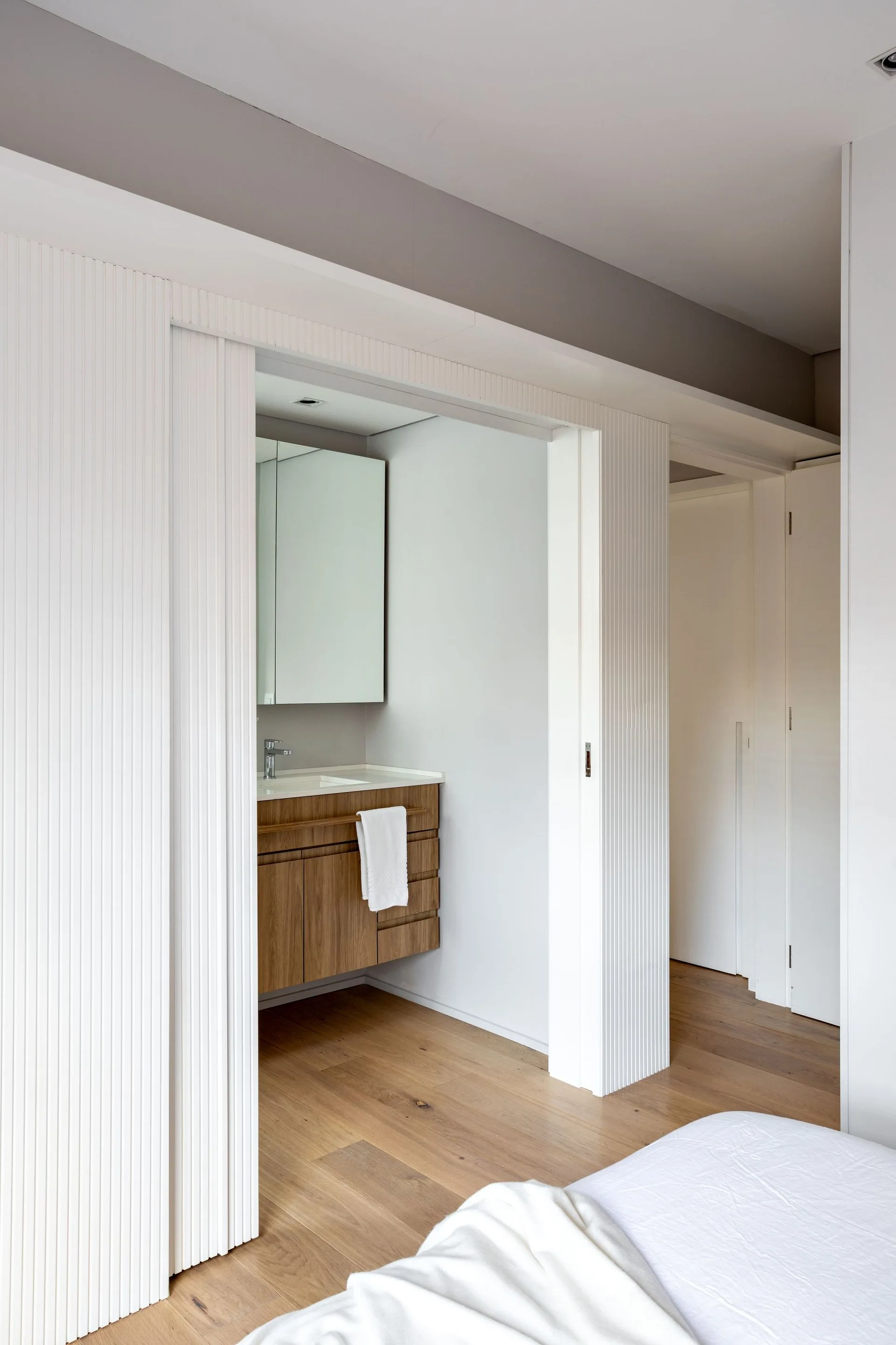 A bedroom view looking into a modern bathroom with white fluted wall panels, a floating wood vanity, and light wood floors.