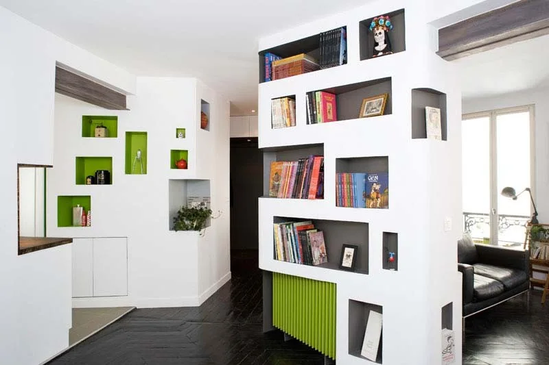 A modern living room features a white custom built-in room divider with colorful recessed shelving niches over a dark wood floor.