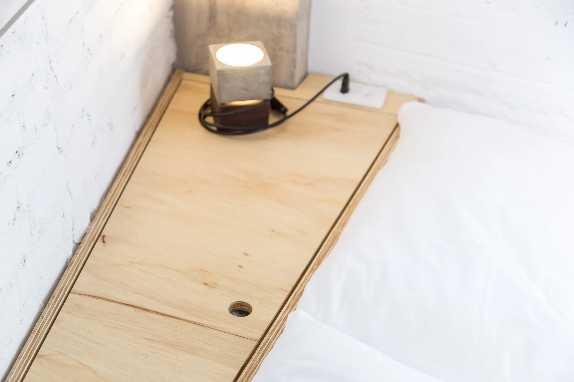 A close-up of an angular plywood bedside shelf with an integrated electrical outlet, a cube lamp, and a white pillow against a painted brick wall.