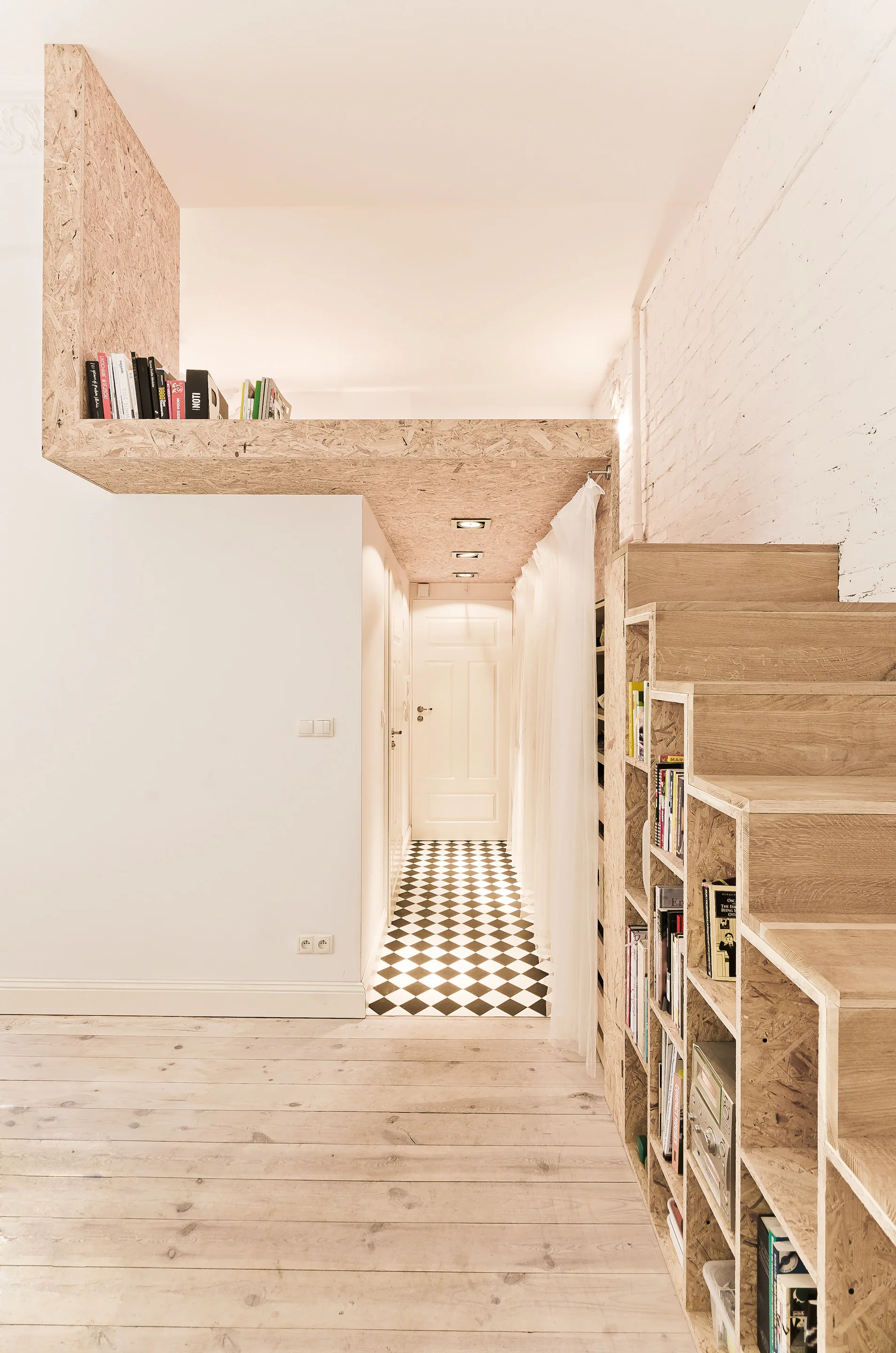 A loft entryway features a wooden staircase with built-in bookshelves, white-washed wood floors, and a black-and-white checkered tile hallway.