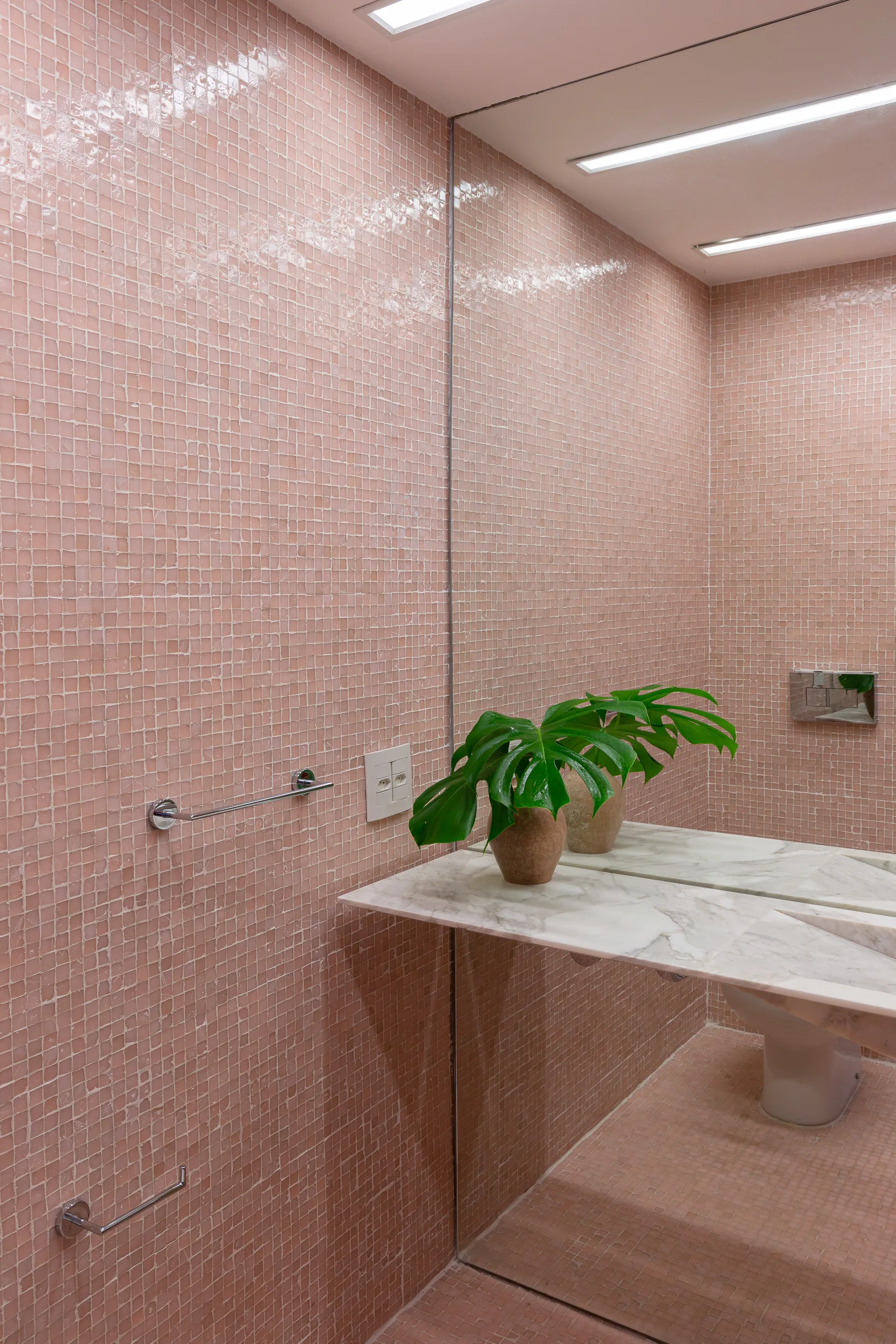 A bathroom with glossy pink mosaic tile walls and floor, a white marble vanity, and a large wall mirror reflecting a potted green plant.