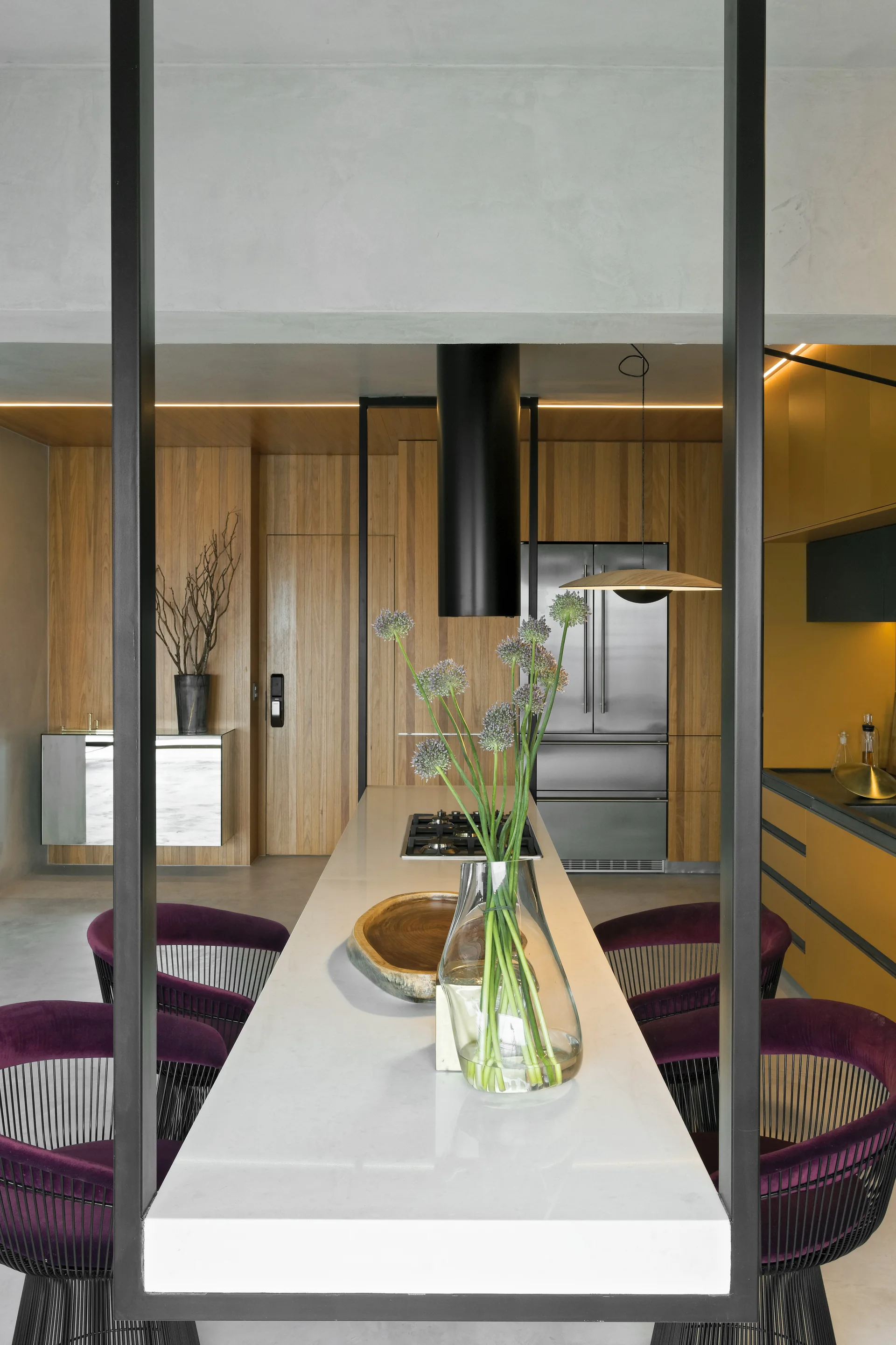 A modern kitchen featuring a long white stone island lined with purple chairs, set against warm wood-paneled walls and a black range hood.