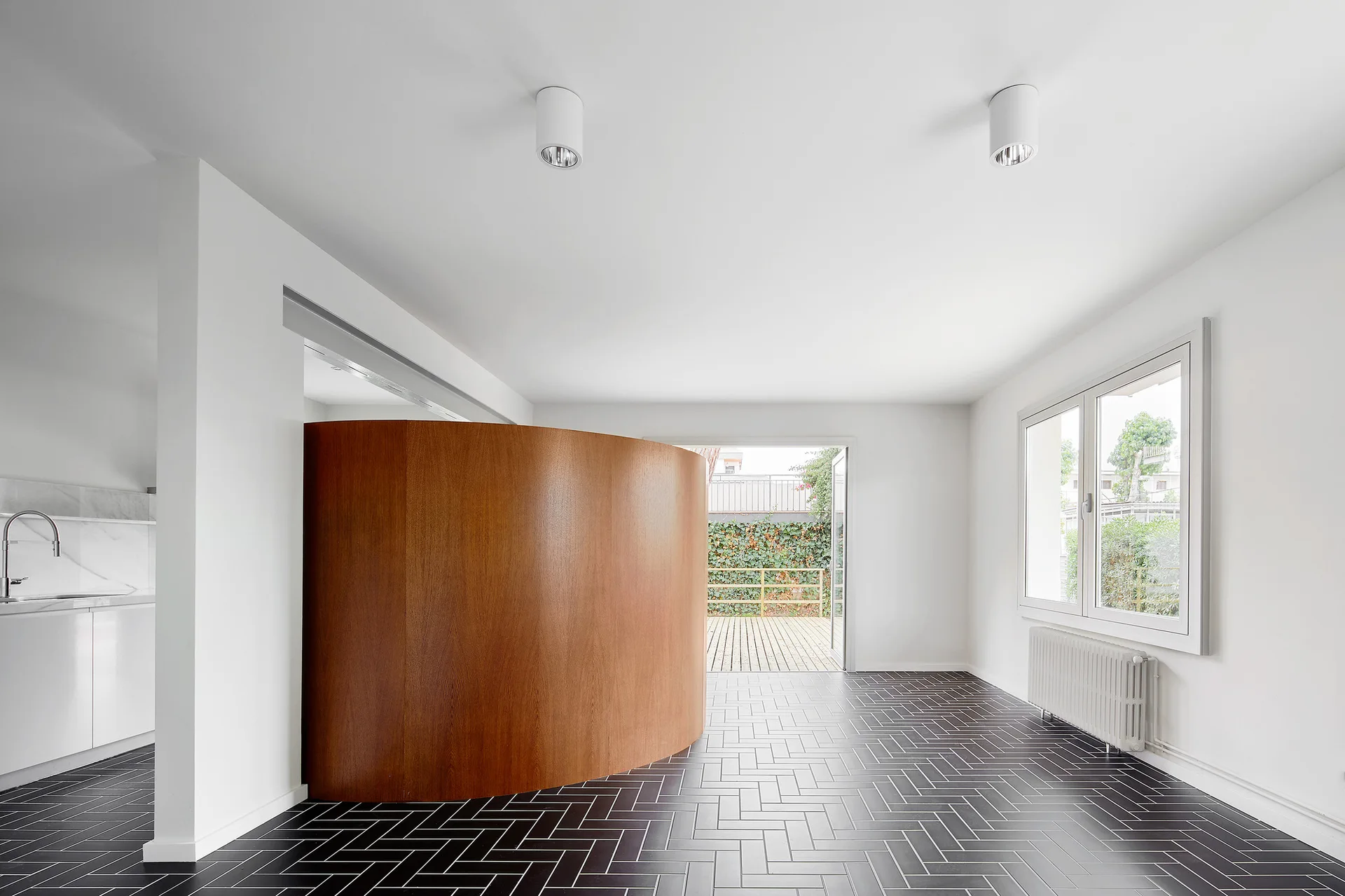 A minimalist living space featuring white walls, black herringbone tile flooring, and a large curved wooden partition wall.