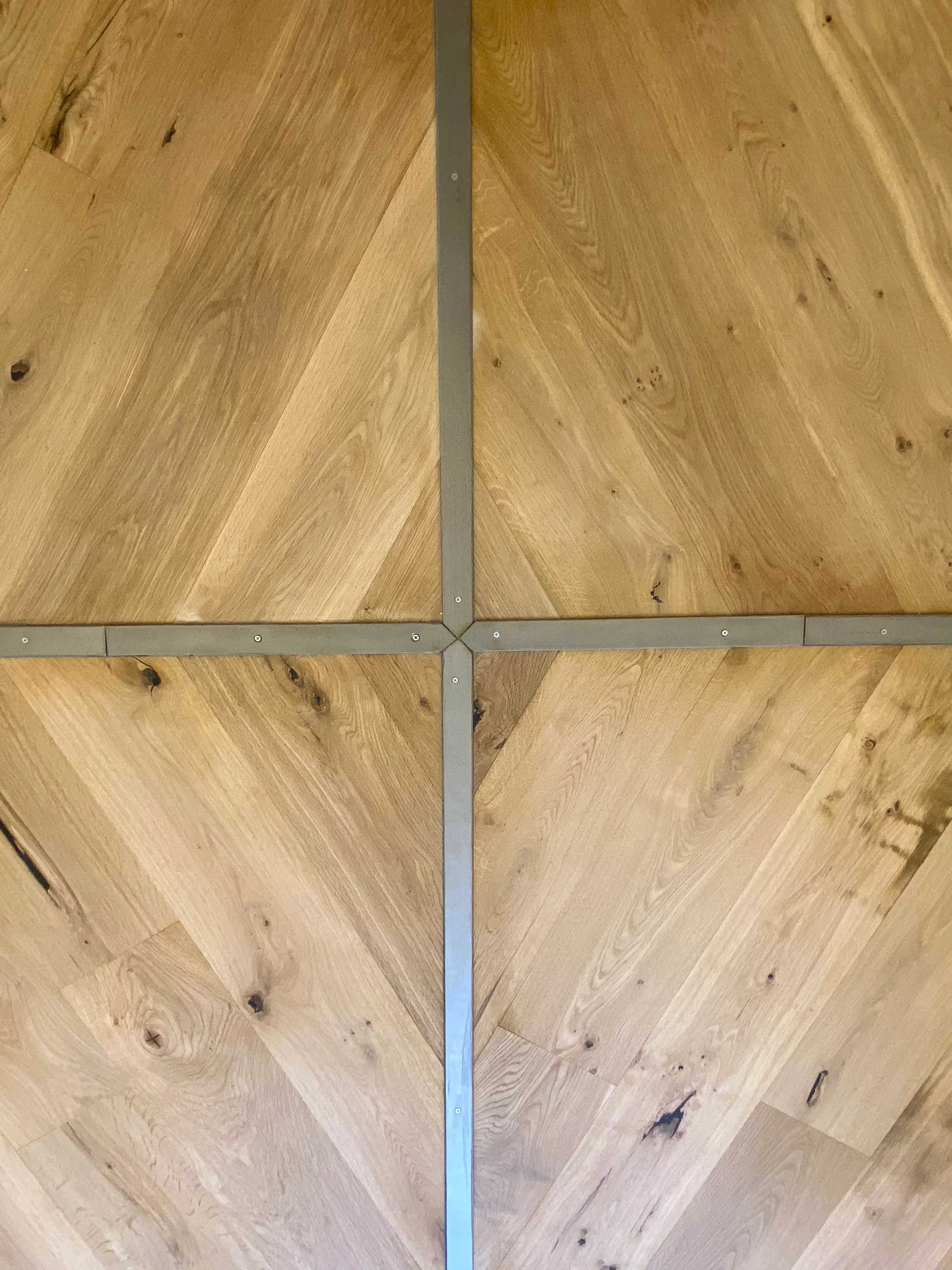 A top-down view of light oak flooring planks arranged in a geometric chevron pattern separated by grey metal inlay strips with visible screws.