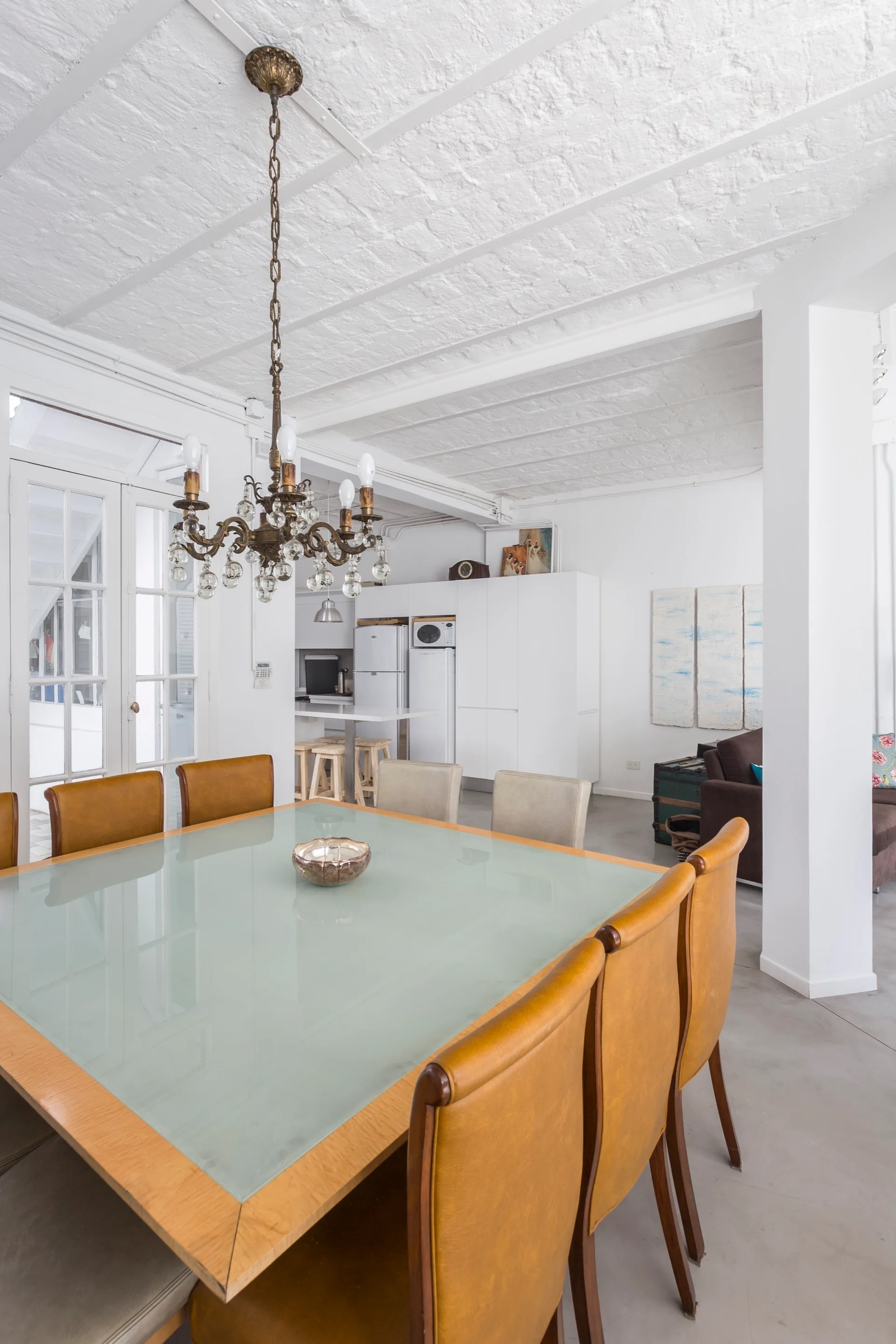 A bright dining room features a glass-top wooden table and tan leather chairs underneath an ornate brass chandelier next to white French doors.