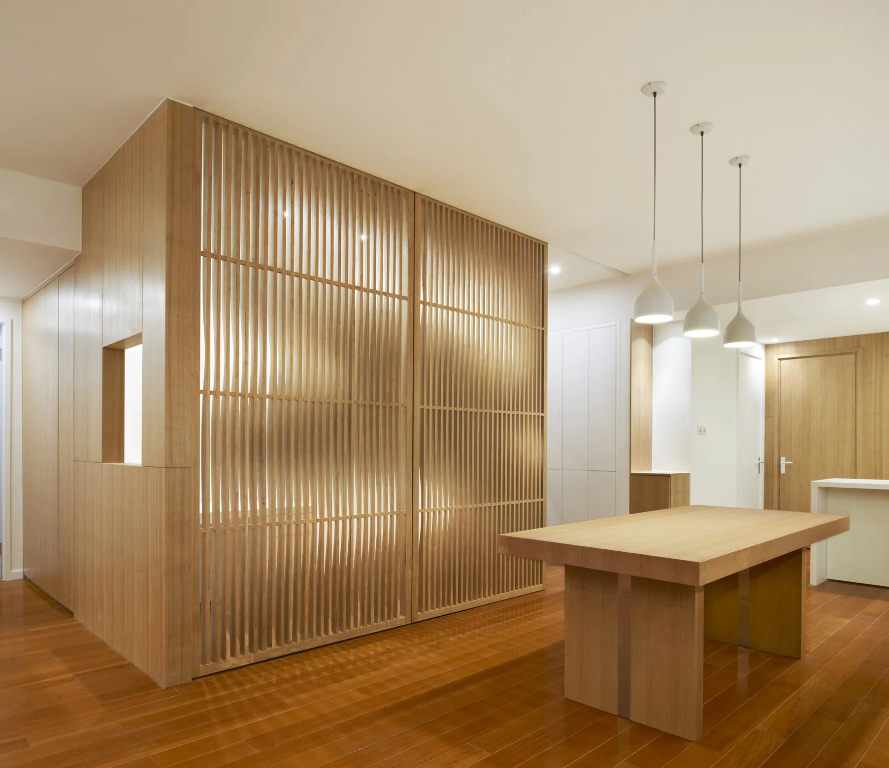 A wooden dining table stands on polished floors beneath three white pendant lights, next to a large, floor-to-ceiling slatted wood partition screen.