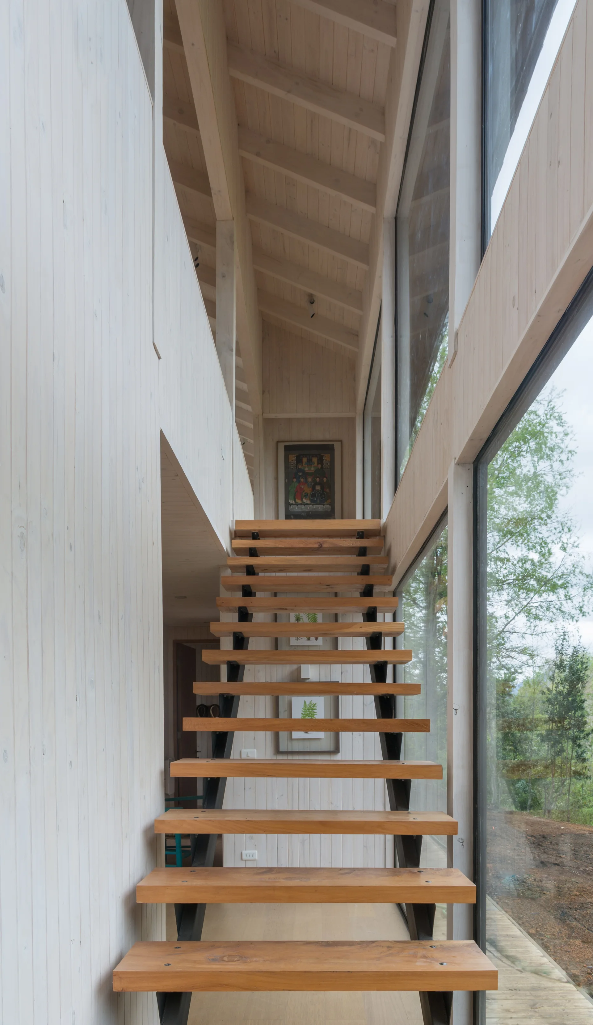 An open-riser wooden staircase sits in a modern hallway with whitewashed wood paneling and a large floor-to-ceiling glass window looking outside.