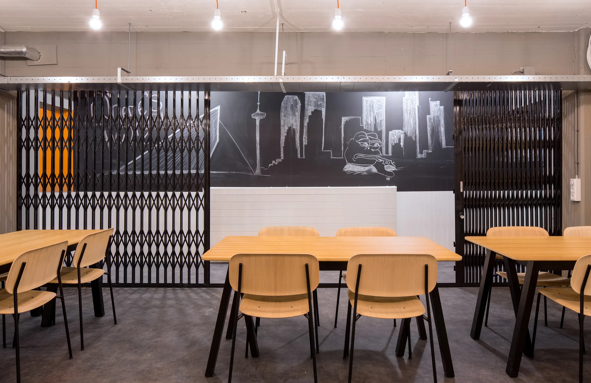 An industrial dining space features light wood tables and chairs arranged in front of a black chalkboard wall and retractable metal security gates.