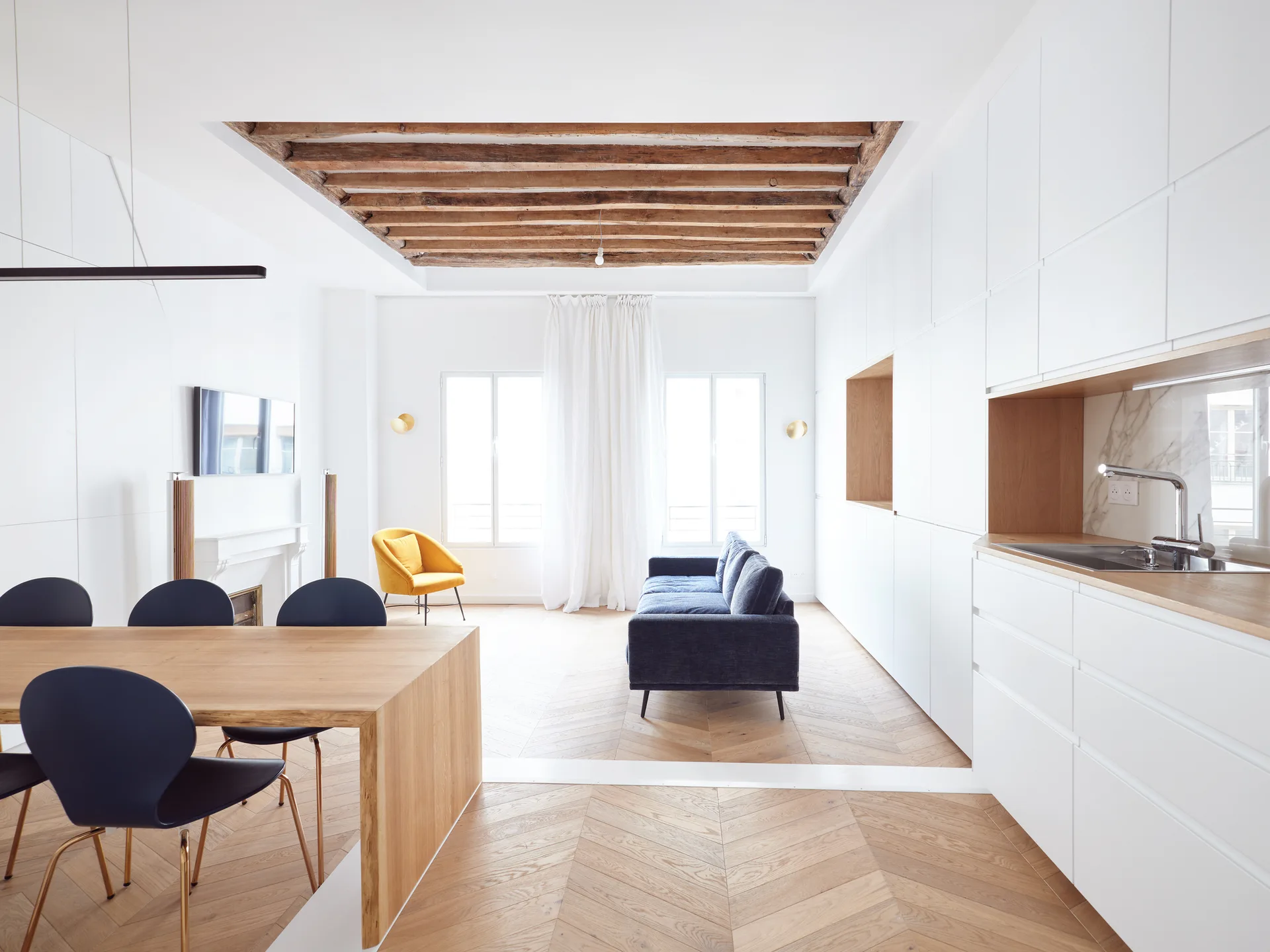 Bright apartment interior showing herringbone wood floors, a recessed timber beam ceiling, and sleek white built-in storage next to a dark sofa.