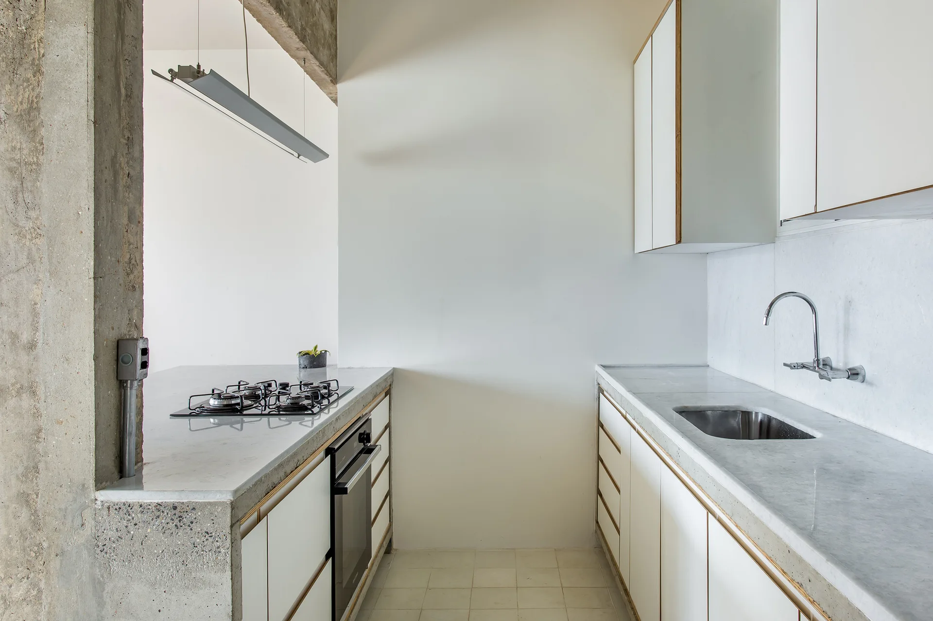 A minimalist galley kitchen features white cabinets with wood trim, light grey stone-like countertops, and an exposed concrete pillar.