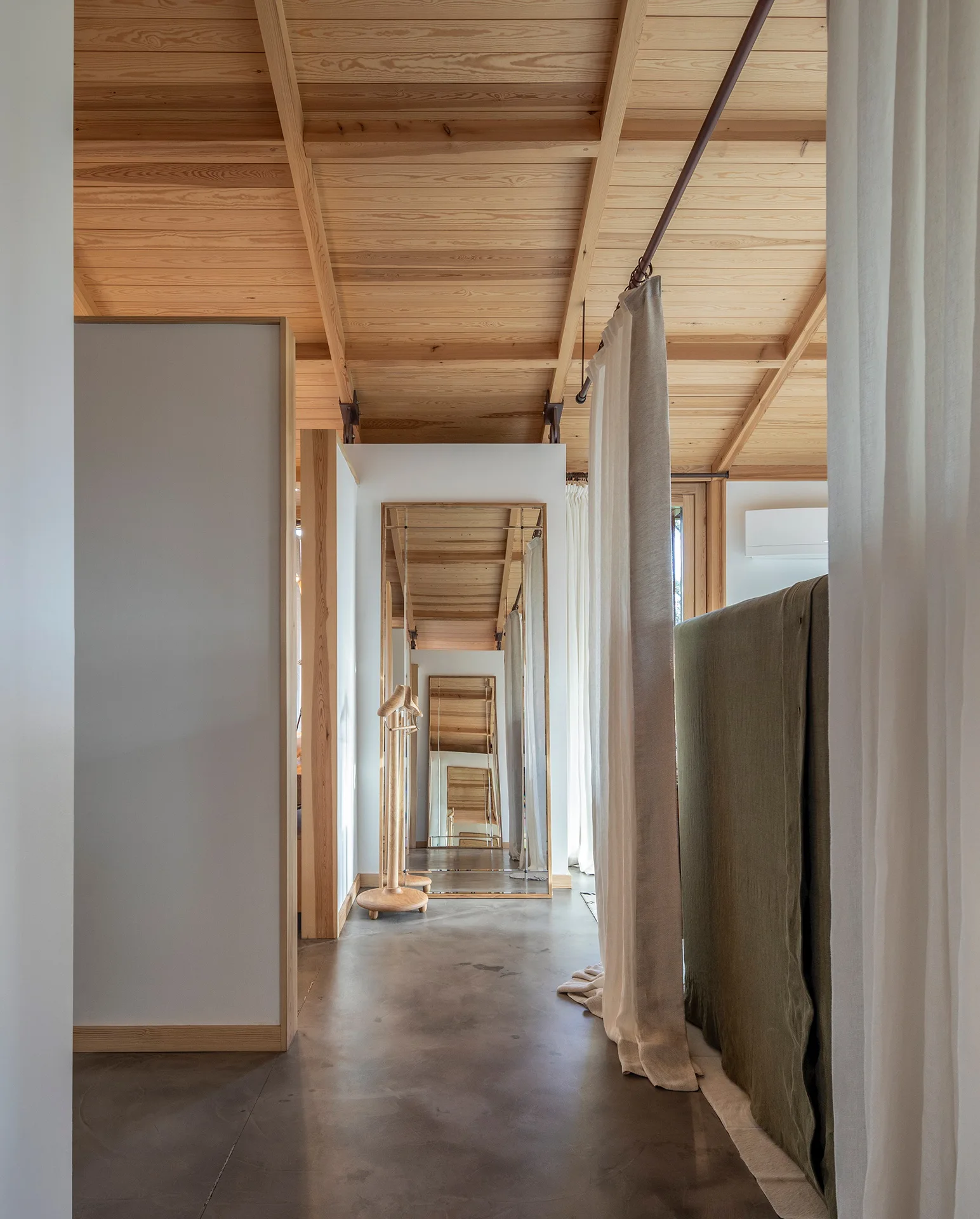 A minimalist hallway features an exposed timber ceiling, grey concrete flooring, a white partition wall, and a large floor mirror reflecting the space.
