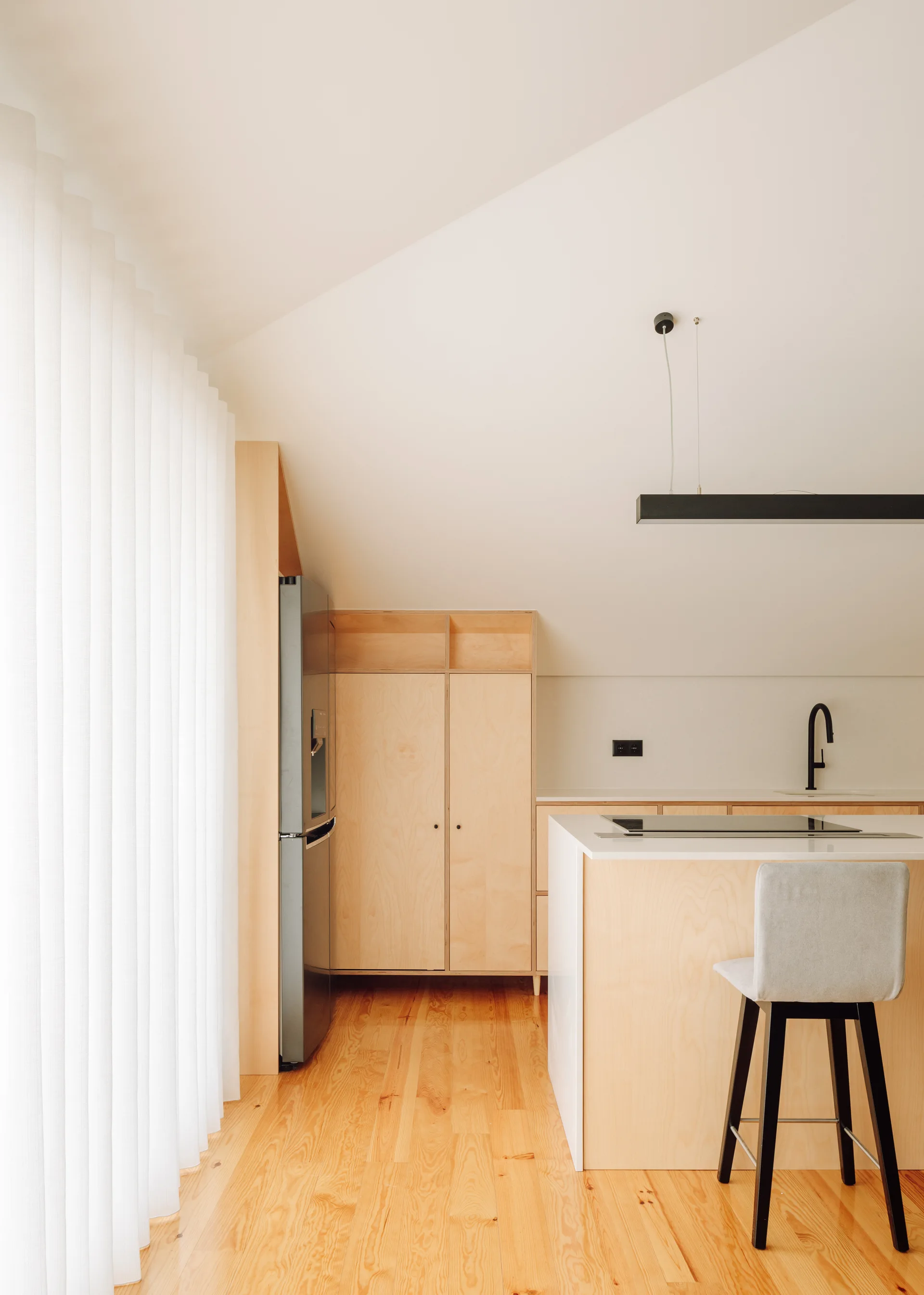 A minimalist kitchen with light plywood cabinets, a sloped ceiling, a white island with a gray barstool, and a black faucet.