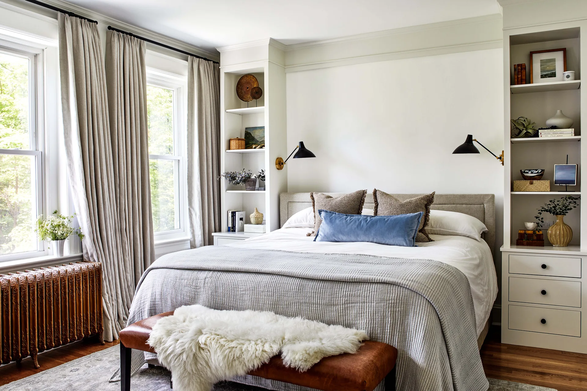 A bedroom with an upholstered bed flanked by white built-in shelving, black wall sconces, and a leather bench with a sheepskin throw.