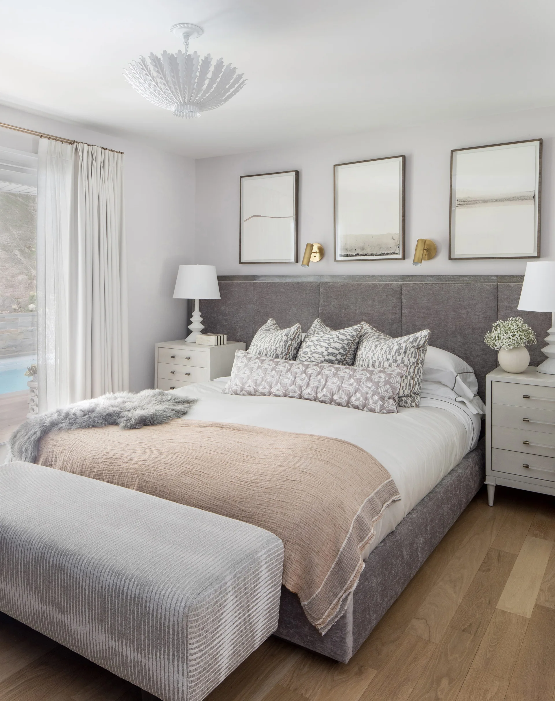 A neutral bedroom featuring a gray upholstered bed frame, off-white nightstands, matching table lamps, and three framed art prints hung symmetrically.