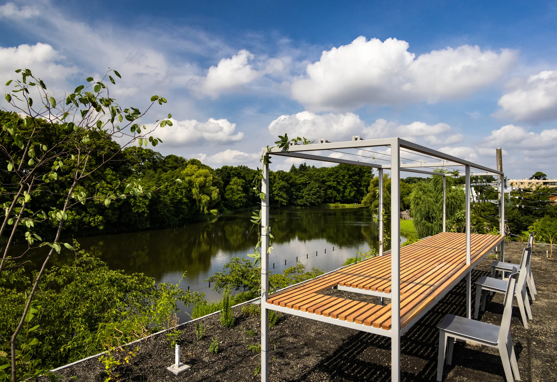 A long slatted wood and metal table with chairs sits on an outdoor gravel terrace overlooking a calm lake surrounded by dense green trees.
