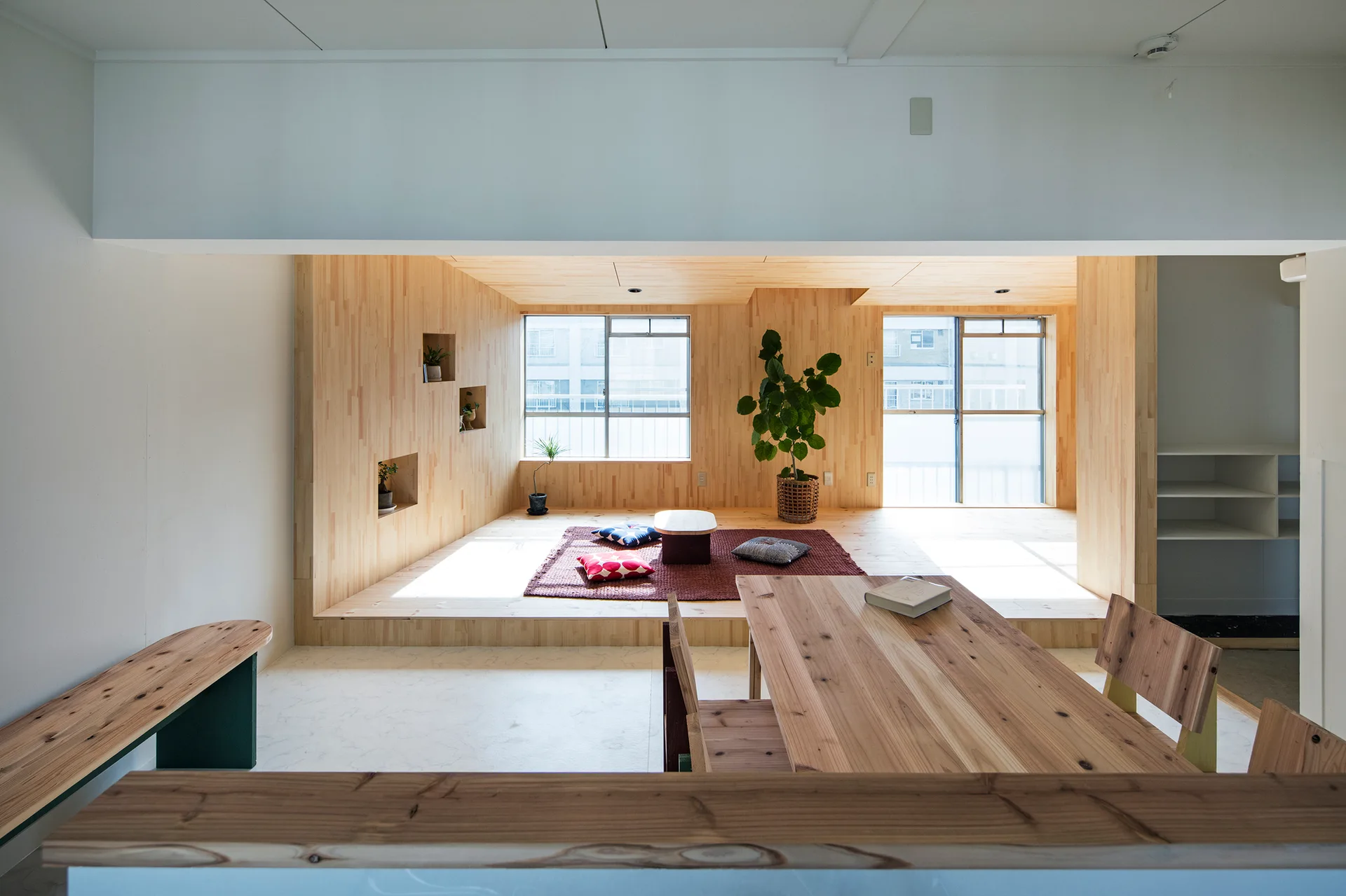 A minimalist dining area with a wooden table sits in front of an elevated wooden living platform with floor cushions, a rug, and large windows.