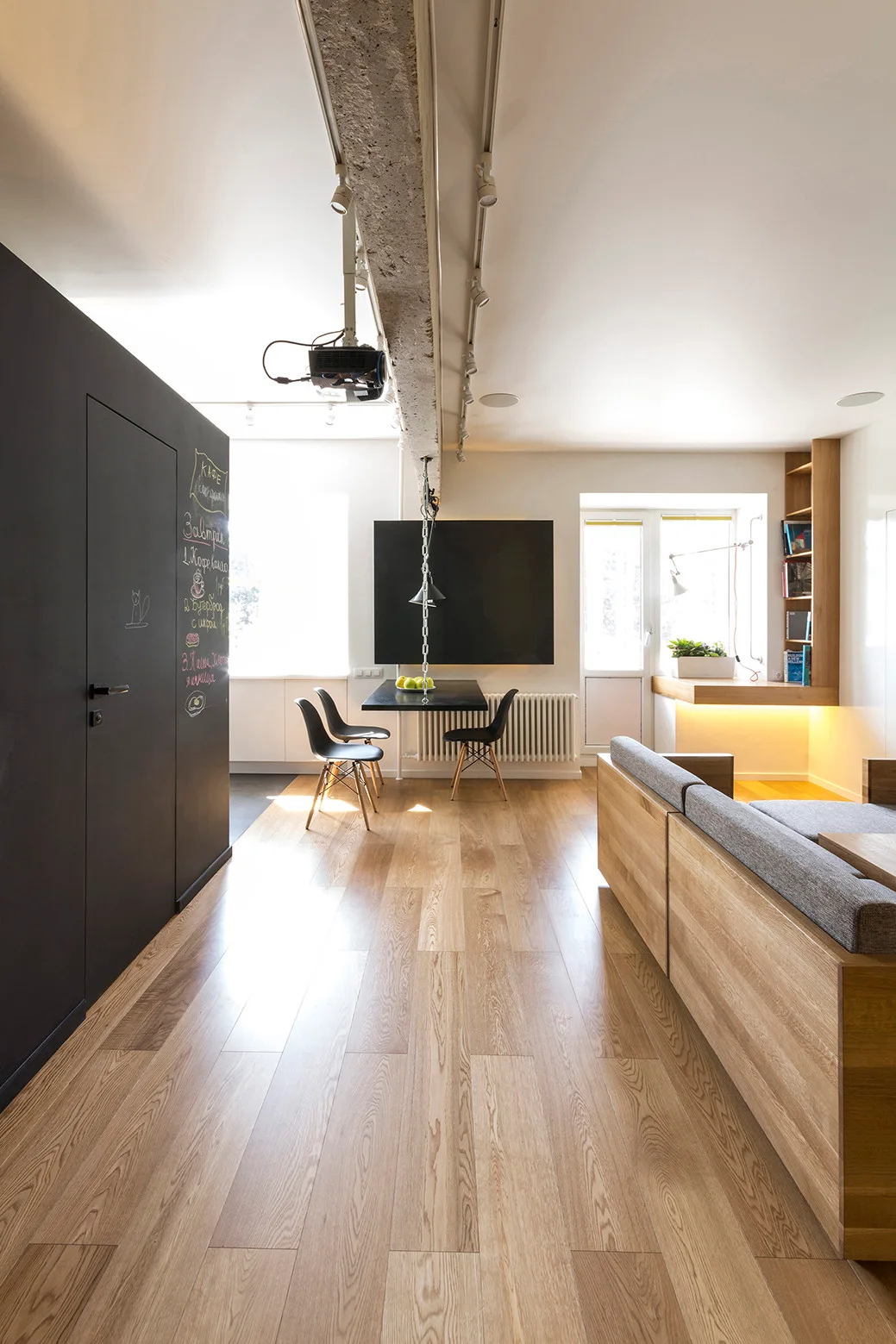 A modern open-plan living room with light wood floors, a concrete ceiling beam, a gray sofa, and a black chalkboard sliding door.