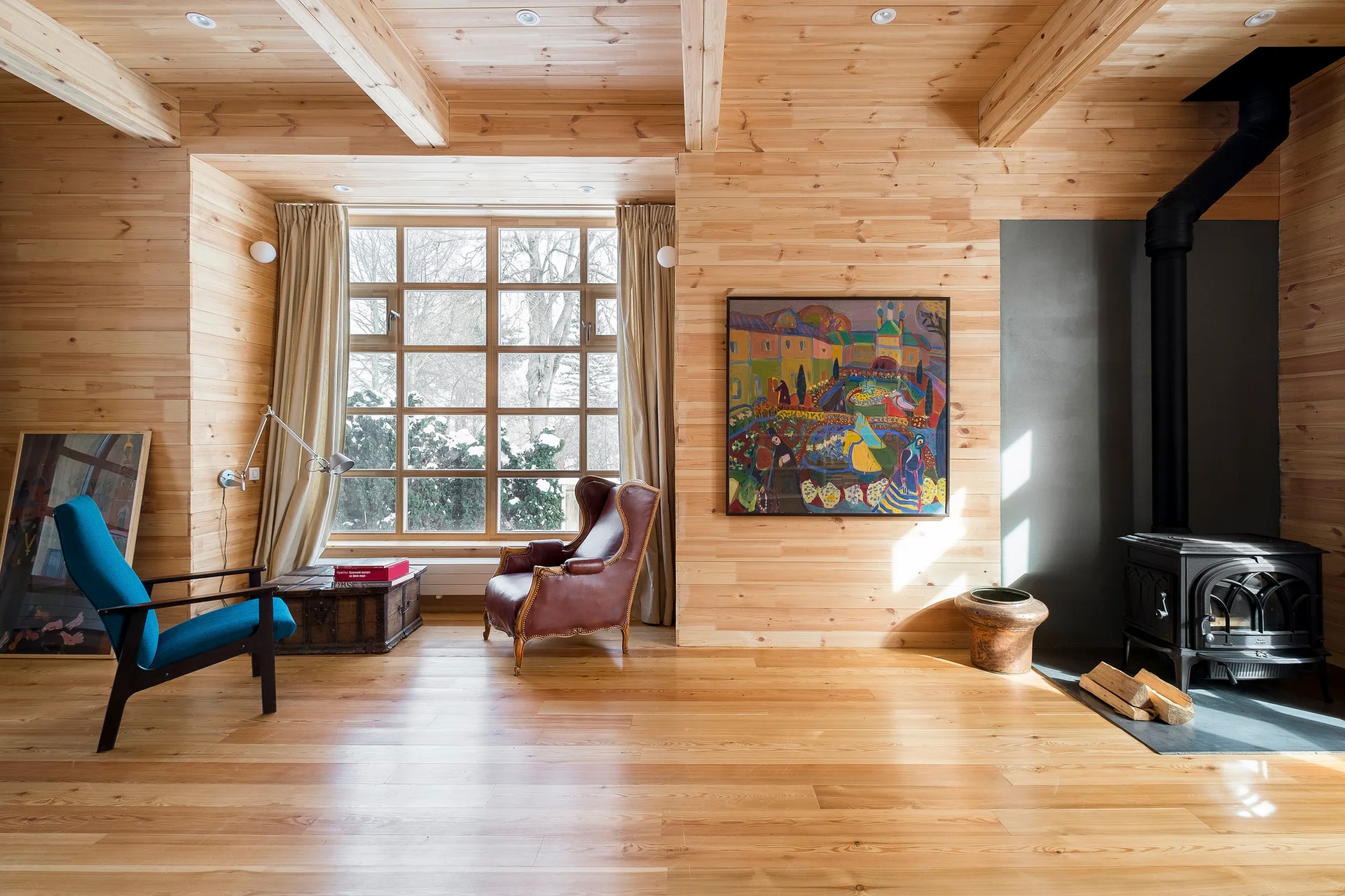 A rustic living room clad in light wood panels features a large window with neutral curtains, a cast iron wood stove, and mismatched armchairs.