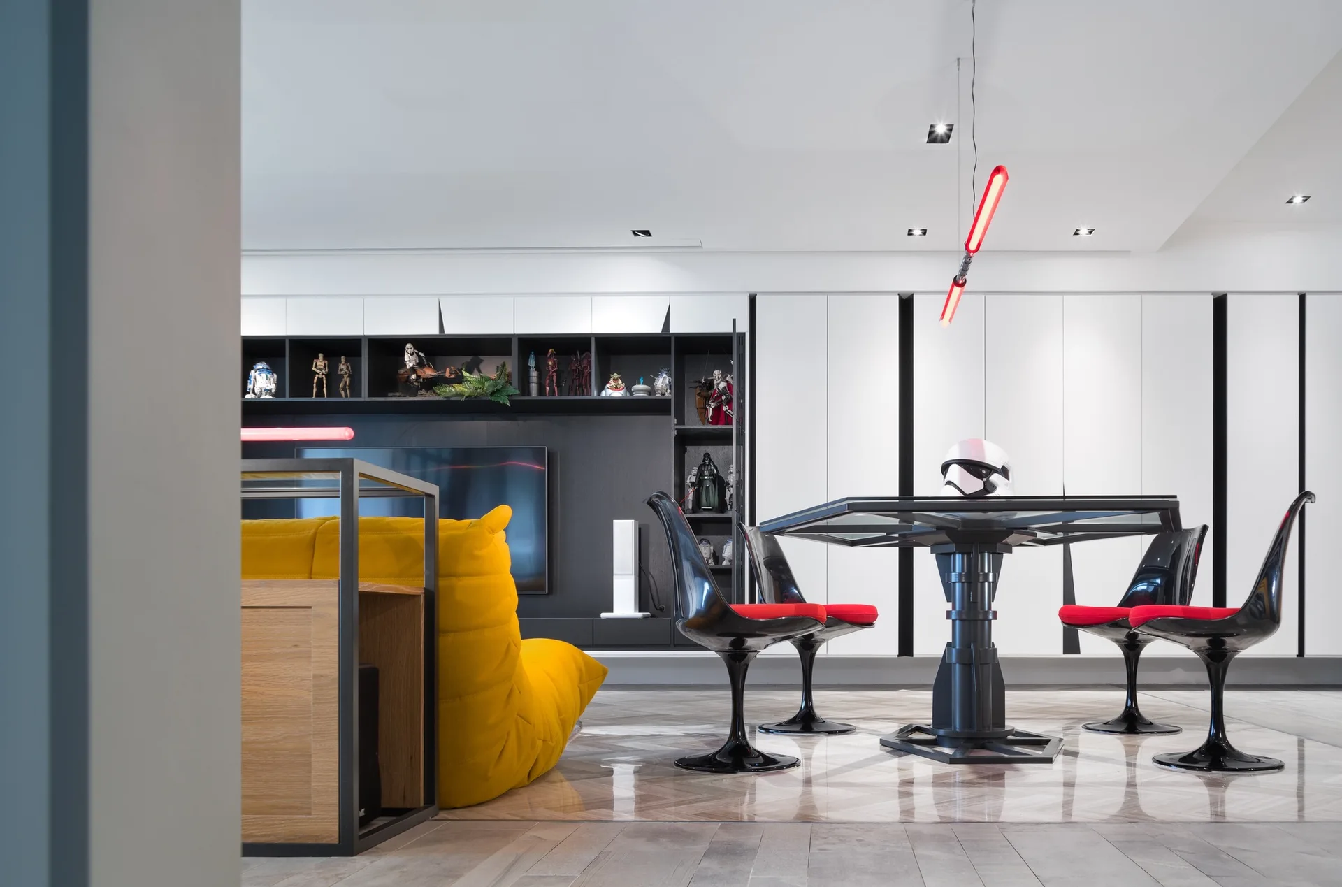 A modern living room features a bright yellow sofa, a black dining set with red-cushioned pedestal chairs, and built-in shelving displaying collectibles.
