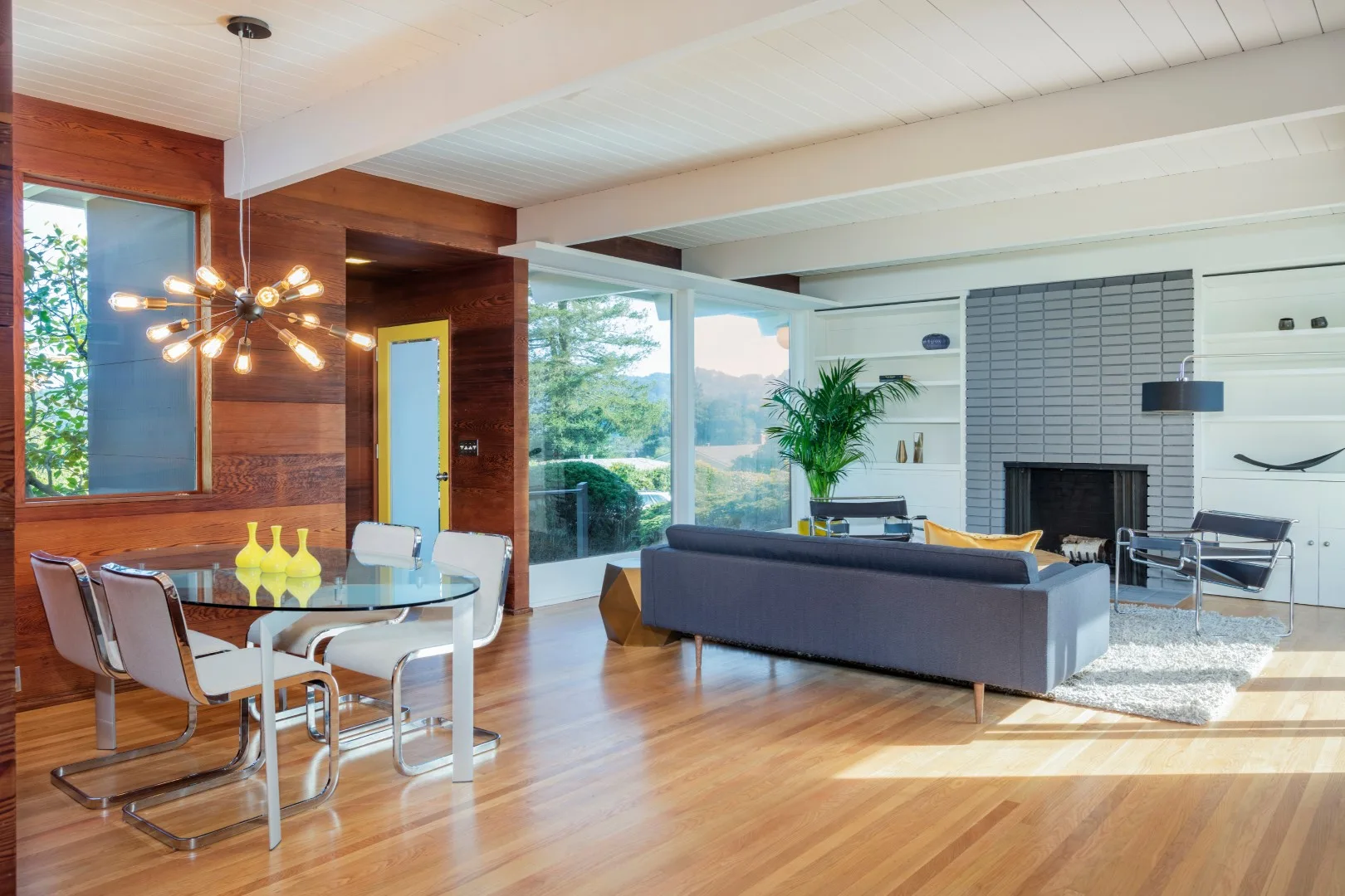 A mid-century modern open living and dining area with horizontal wood wall paneling, a grey brick fireplace, and a glass dining table.