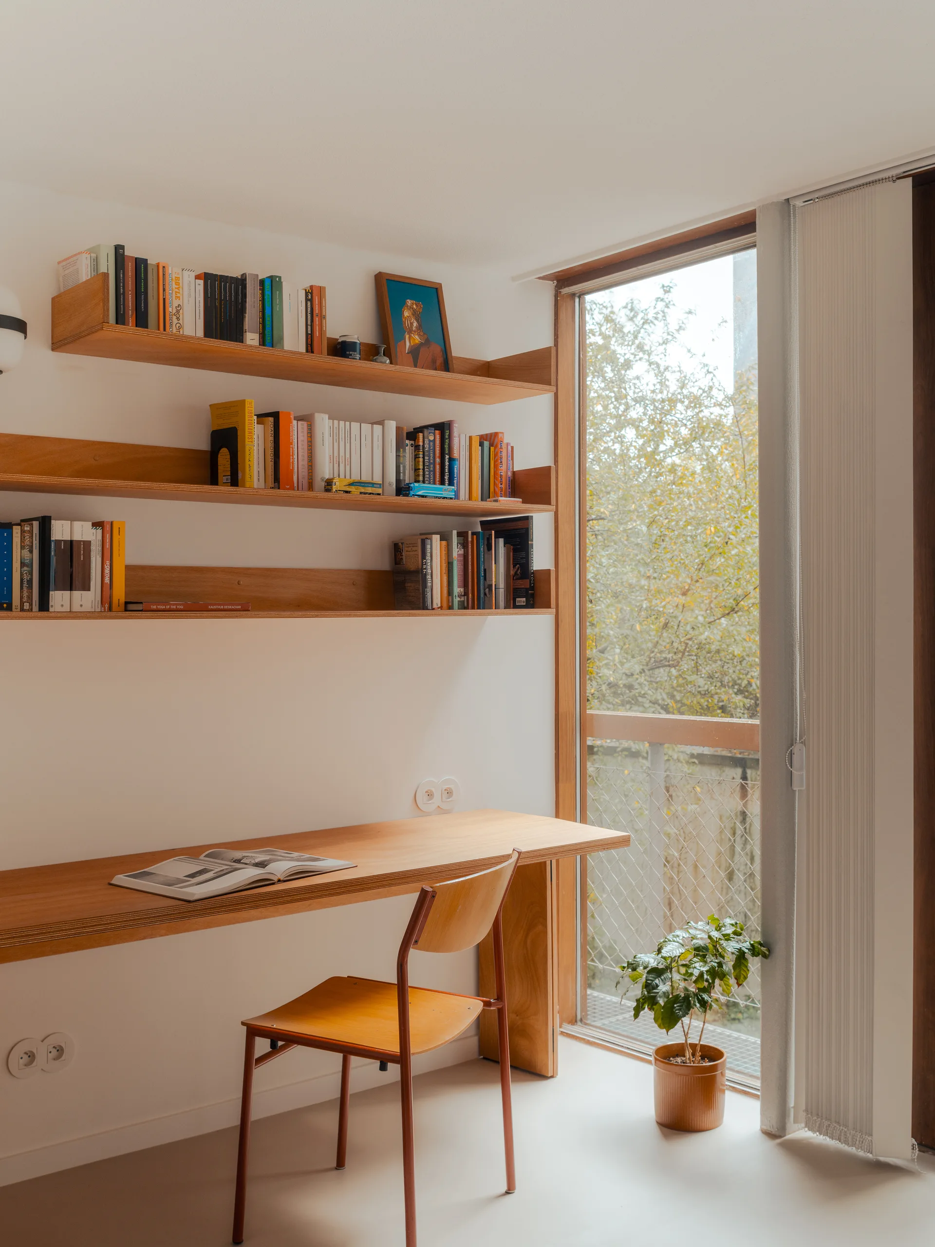 A minimalist home office features a wall-mounted wooden desk, matching floating shelves filled with books, a wooden chair, and a large window.