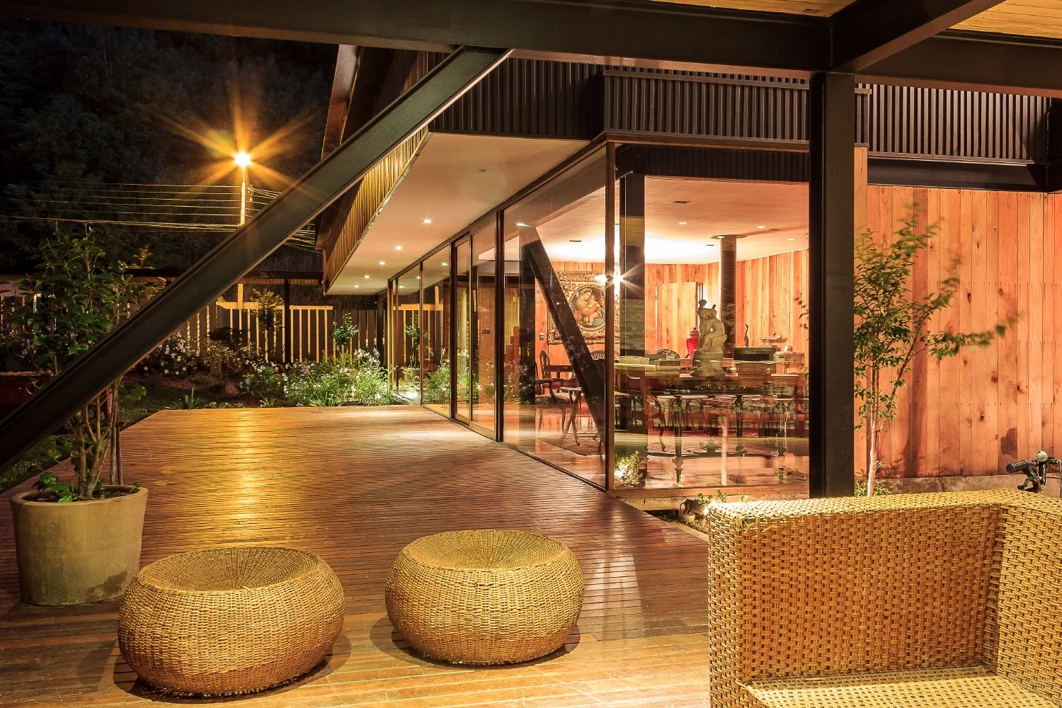 A spacious wooden deck at night featuring round wicker ottomans, a matching wicker sofa, and a large glass wall displaying an illuminated interior.