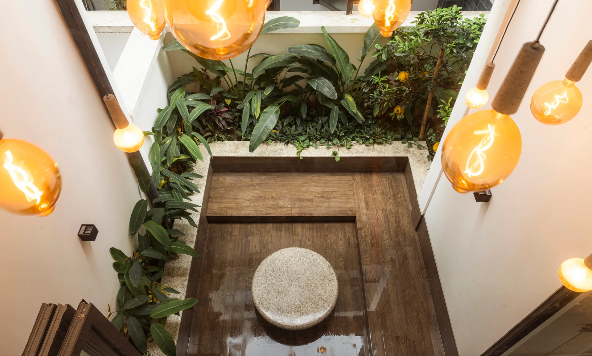 An overhead view of an interior courtyard with tiered dark wood decking, a circular stone stool, lush tropical plants, and hanging amber light bulbs.