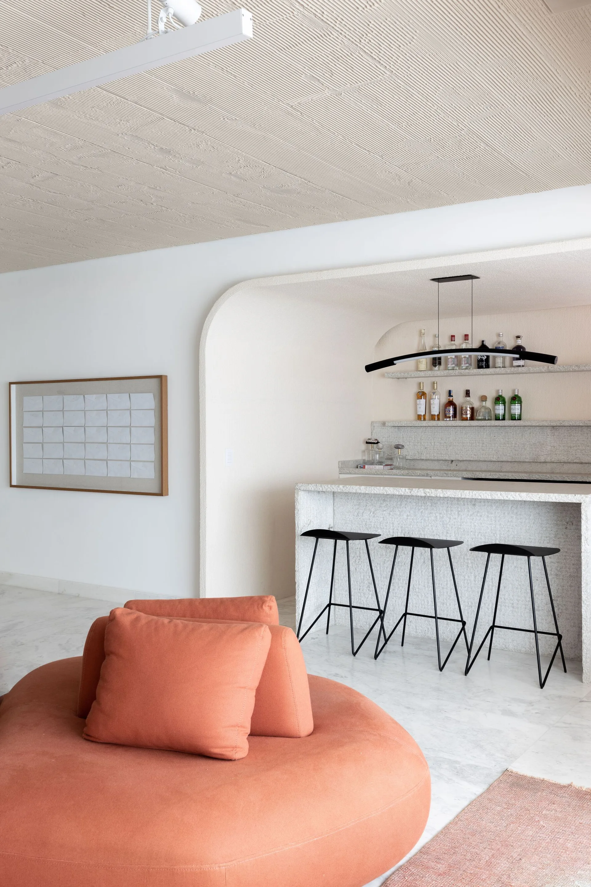 A minimalist home bar featuring a textured stone counter, black metal stools, and a plaster archway, seen past a round terracotta-colored lounge sofa.