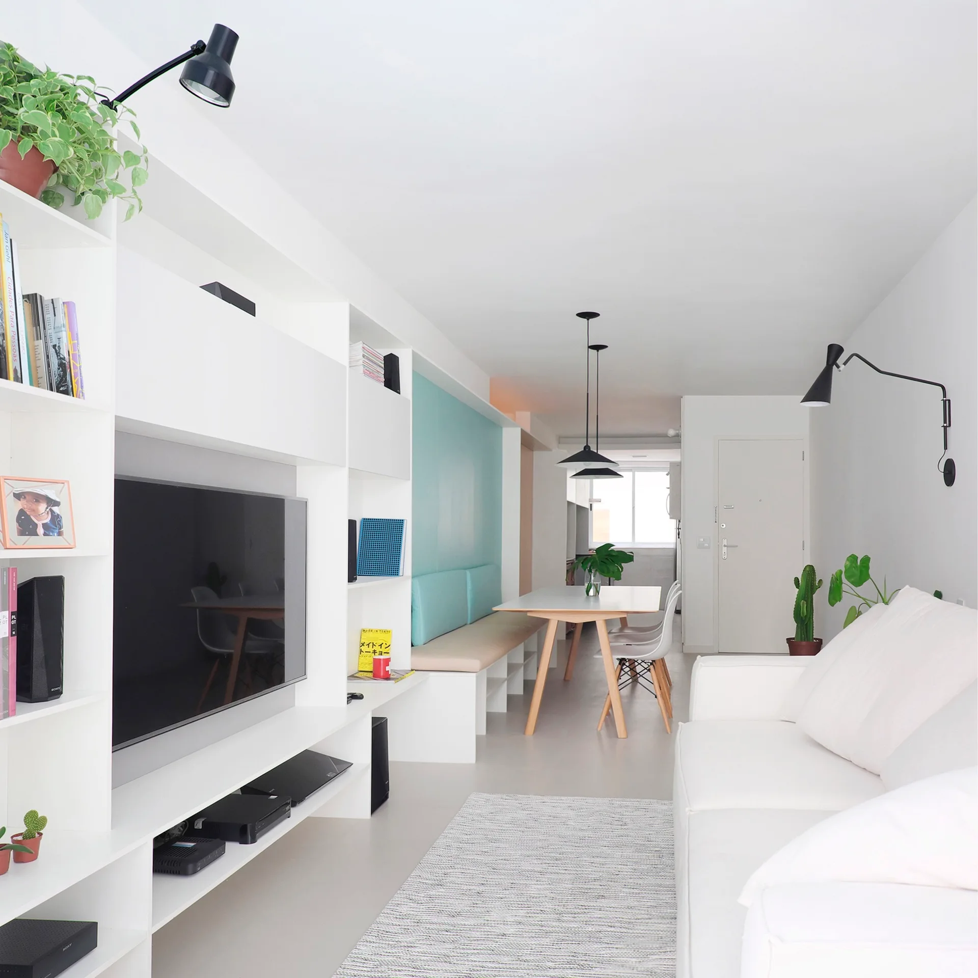 A bright white minimalist living room features a custom media wall, a white sofa, and a dining area with a light blue accented built-in bench.
