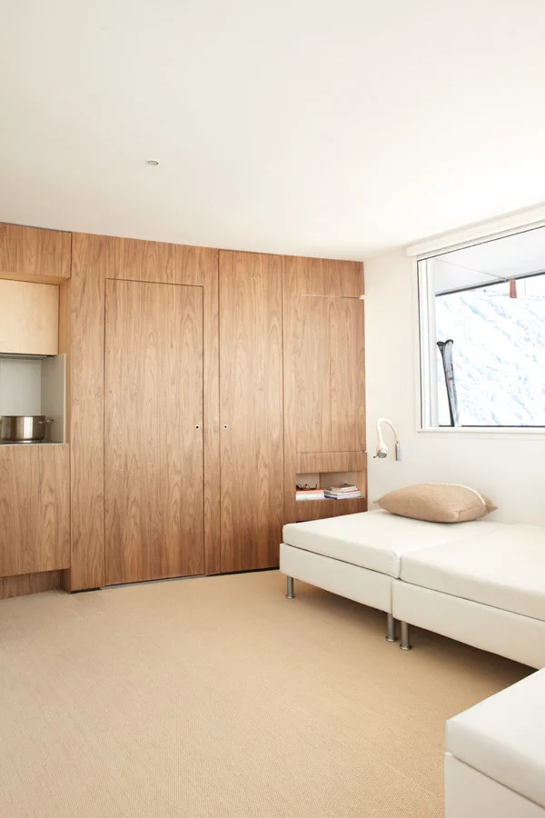A minimalist bedroom features a floor-to-ceiling wood wardrobe wall, a low white daybed, and a large window showcasing a snowy outdoor view.