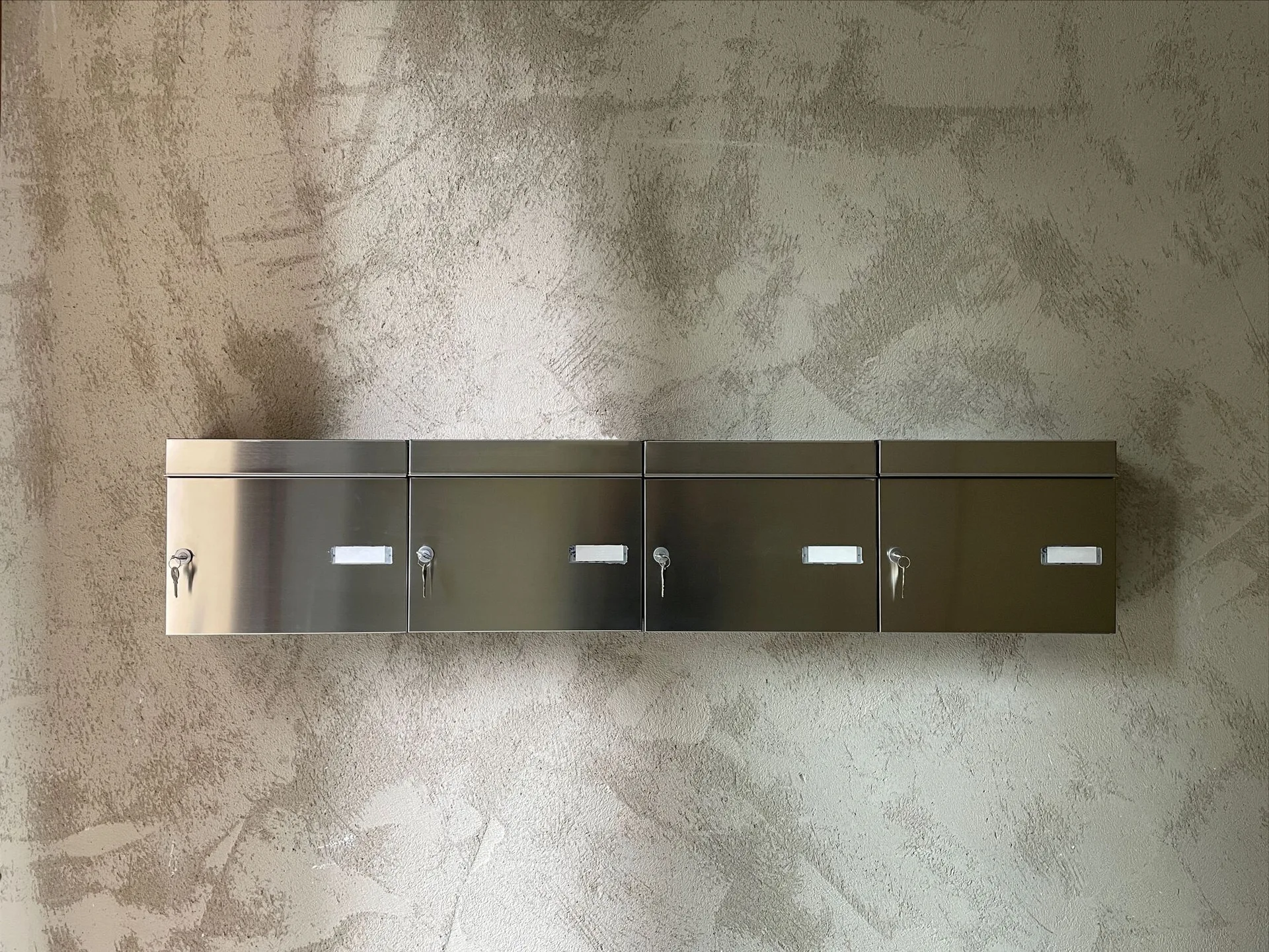Four wall-mounted brushed metal mailboxes with keys inserted are aligned on a textured grey plaster wall.