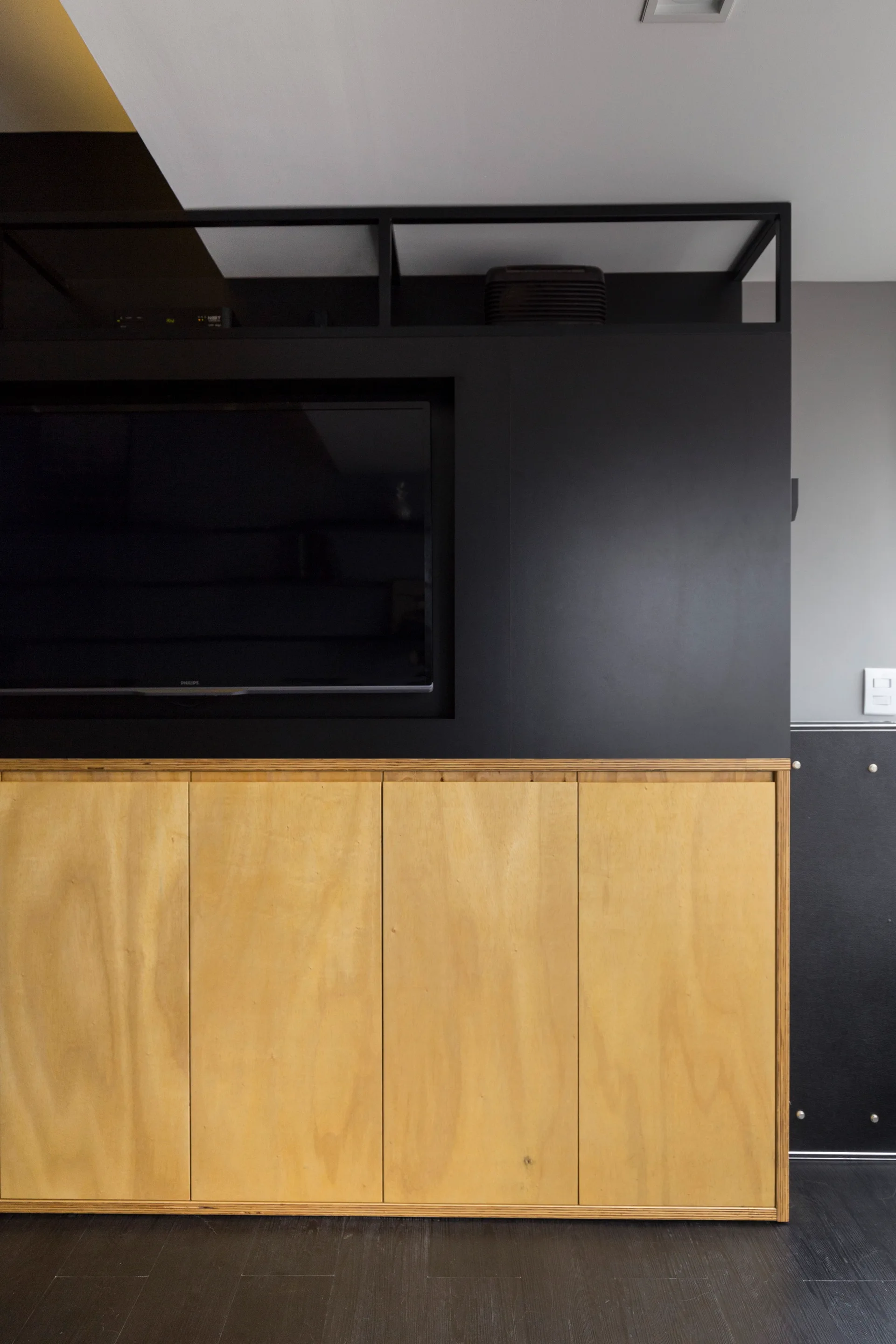 A minimalist built-in media console with light plywood lower cabinets and a recessed television in a matte black upper section.