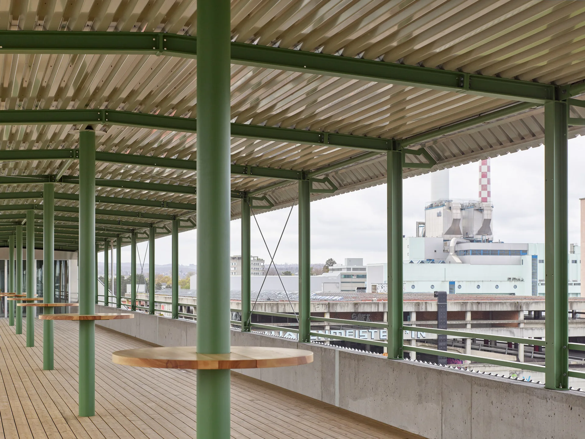 An outdoor covered terrace with wood decking, green painted columns with attached circular wooden tables, and a corrugated metal roof.