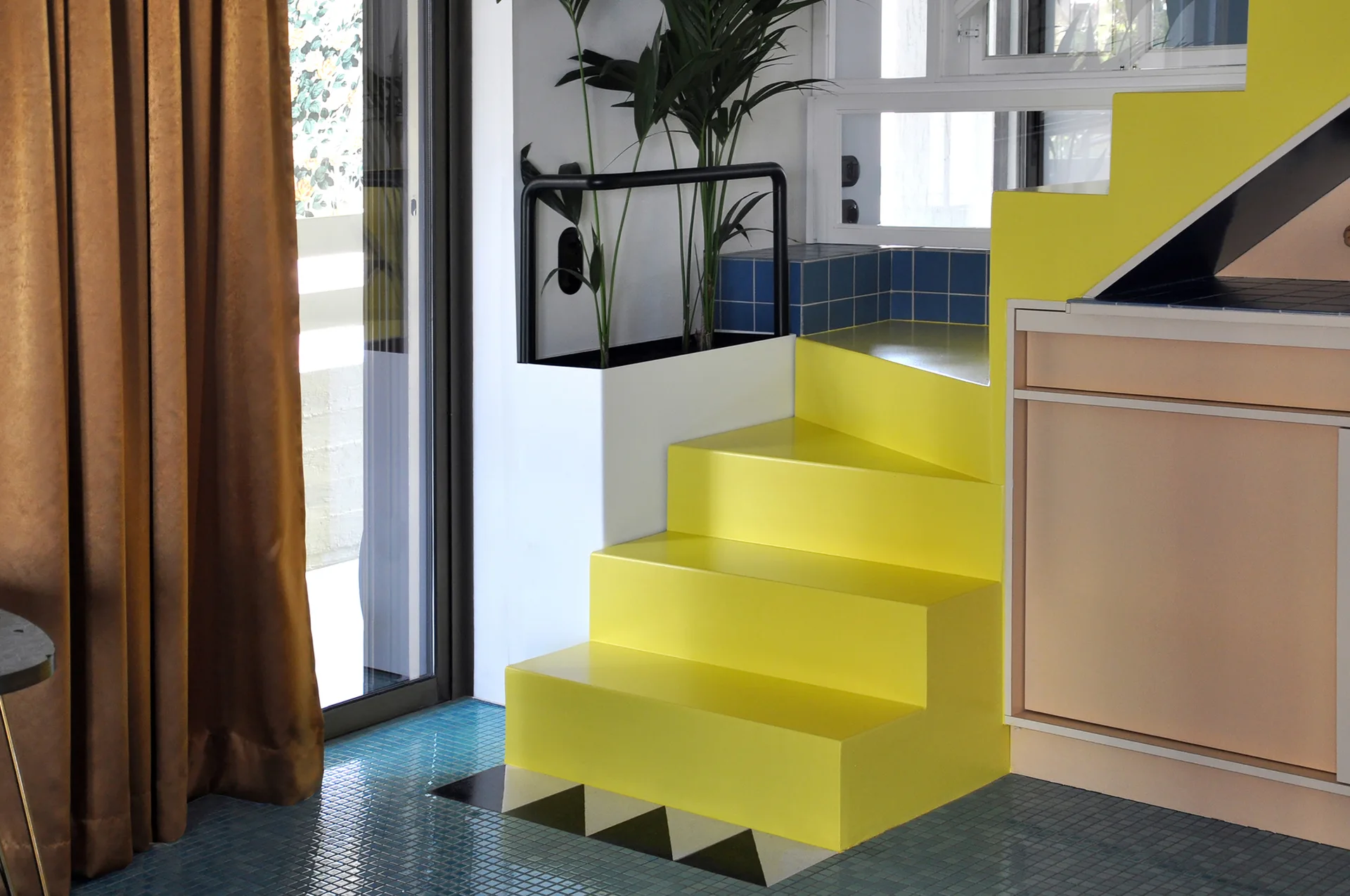 A bright yellow staircase with a black handrail sits next to a peach cabinet and green plants on a blue mosaic tiled floor.