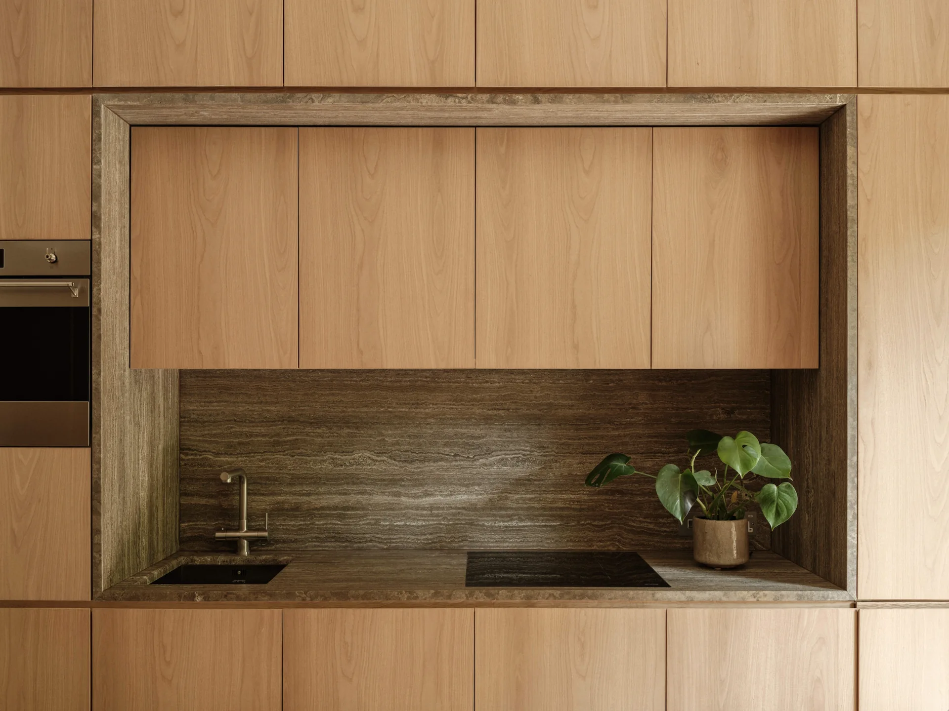 Modern kitchen features flat-panel wood cabinetry with a recessed alcove lined in veined brown stone containing a sink, cooktop, and small plant.