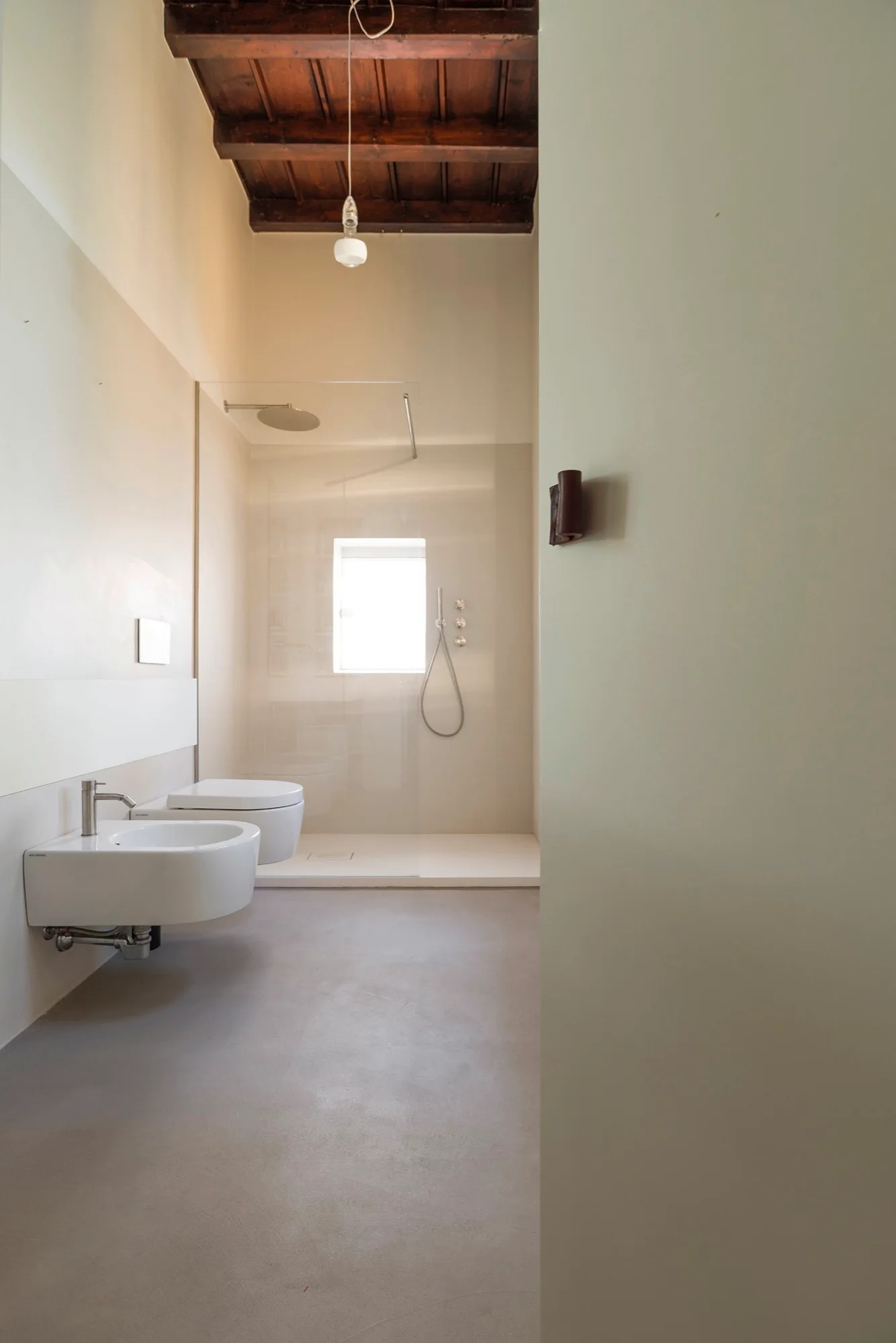 A minimalist bathroom featuring a wall-mounted bidet and toilet, a glass walk-in shower with a window, and an exposed dark wood beamed ceiling.