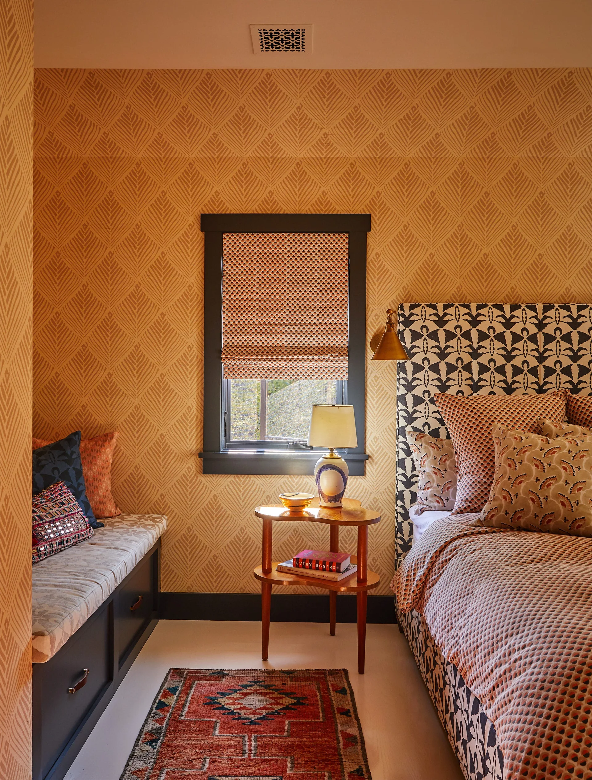 A bedroom features golden leaf-patterned wallpaper, a dark built-in bench, a wooden side table, and a bed with a graphic patterned headboard.