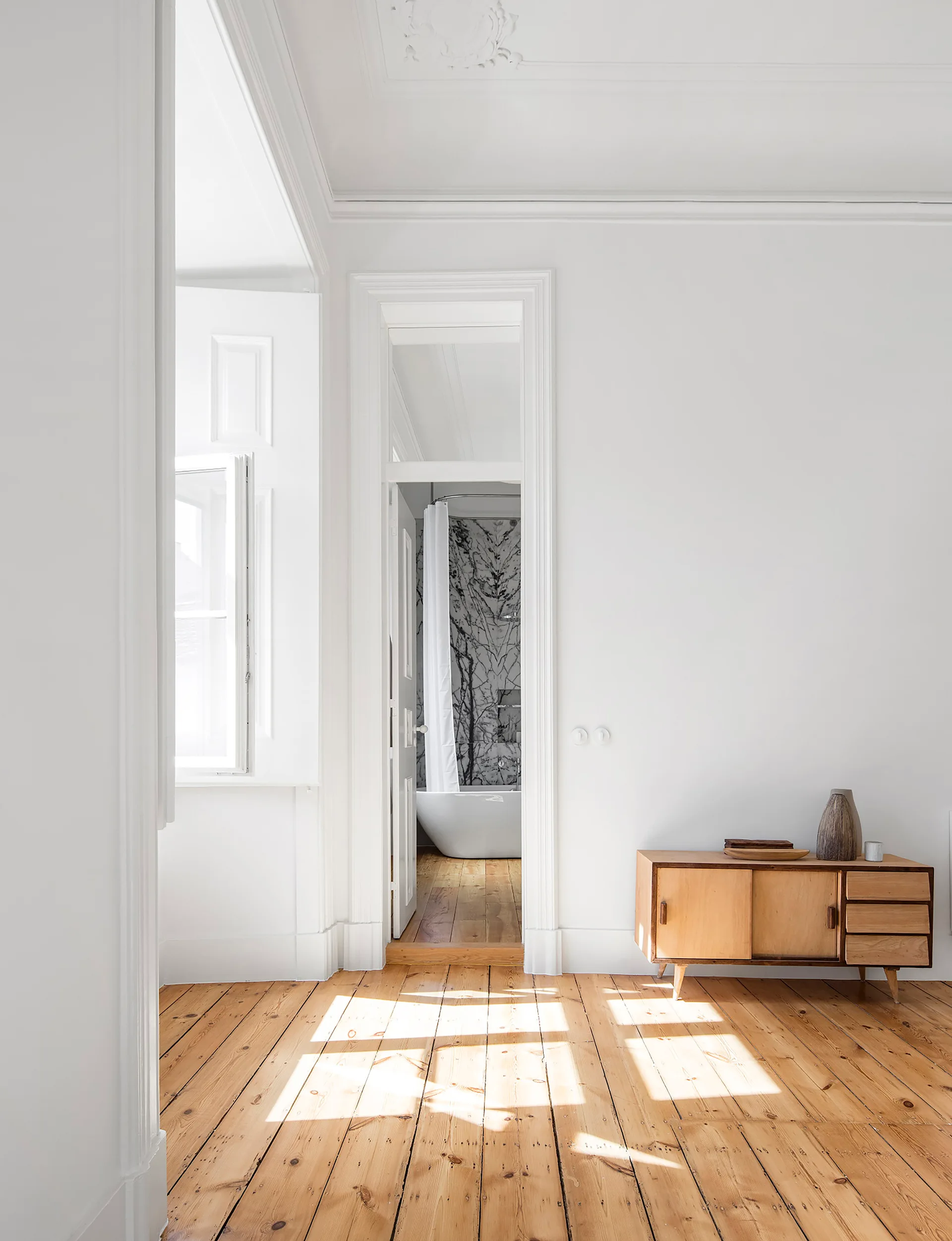 A sunlit white room with wood floors and a wooden sideboard opens to a bathroom with a freestanding tub and veined marble wall.