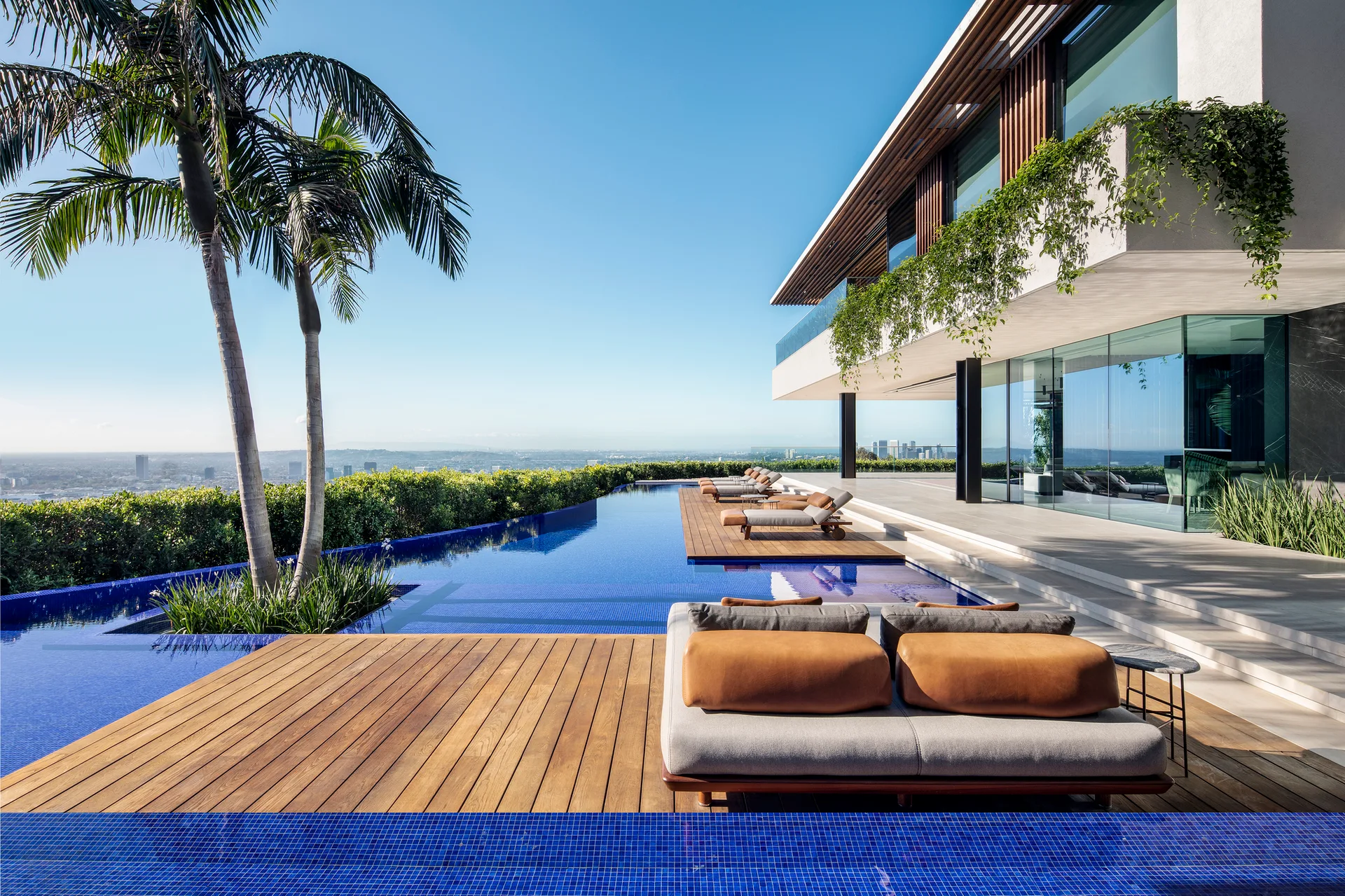 A modern infinity pool with wooden deck platforms, tan lounge furniture, and a palm tree next to a white villa under a clear blue sky.