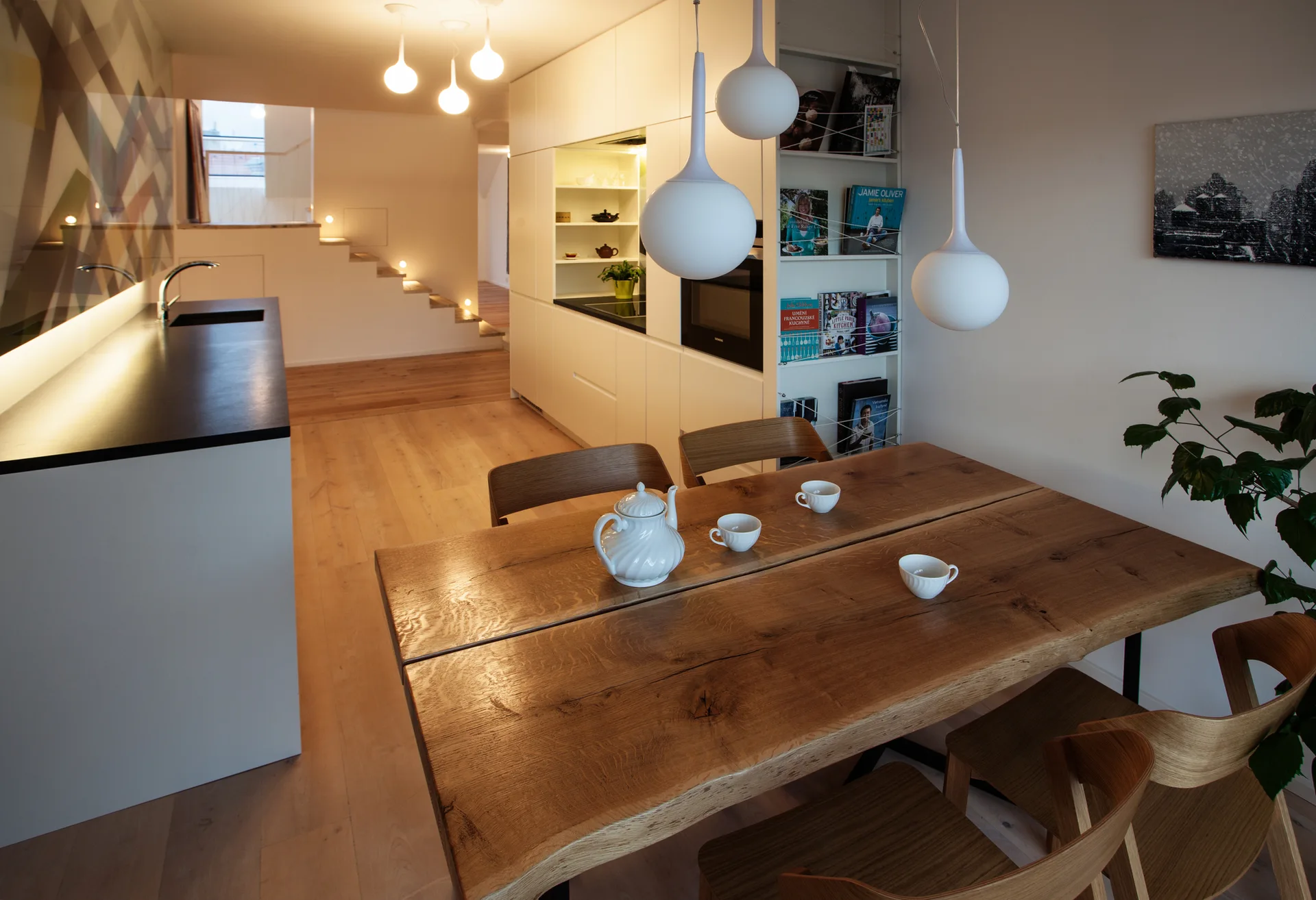 A live-edge wood dining table with a ceramic tea set sits in an open-plan kitchen featuring minimalist white cabinets and spherical pendant lights.
