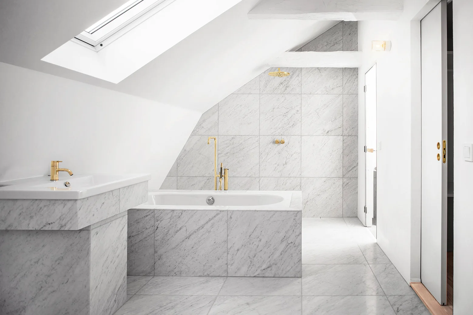 An attic bathroom featuring a sloped ceiling with a skylight, a marble-covered bathtub and vanity, and metallic gold plumbing fixtures.