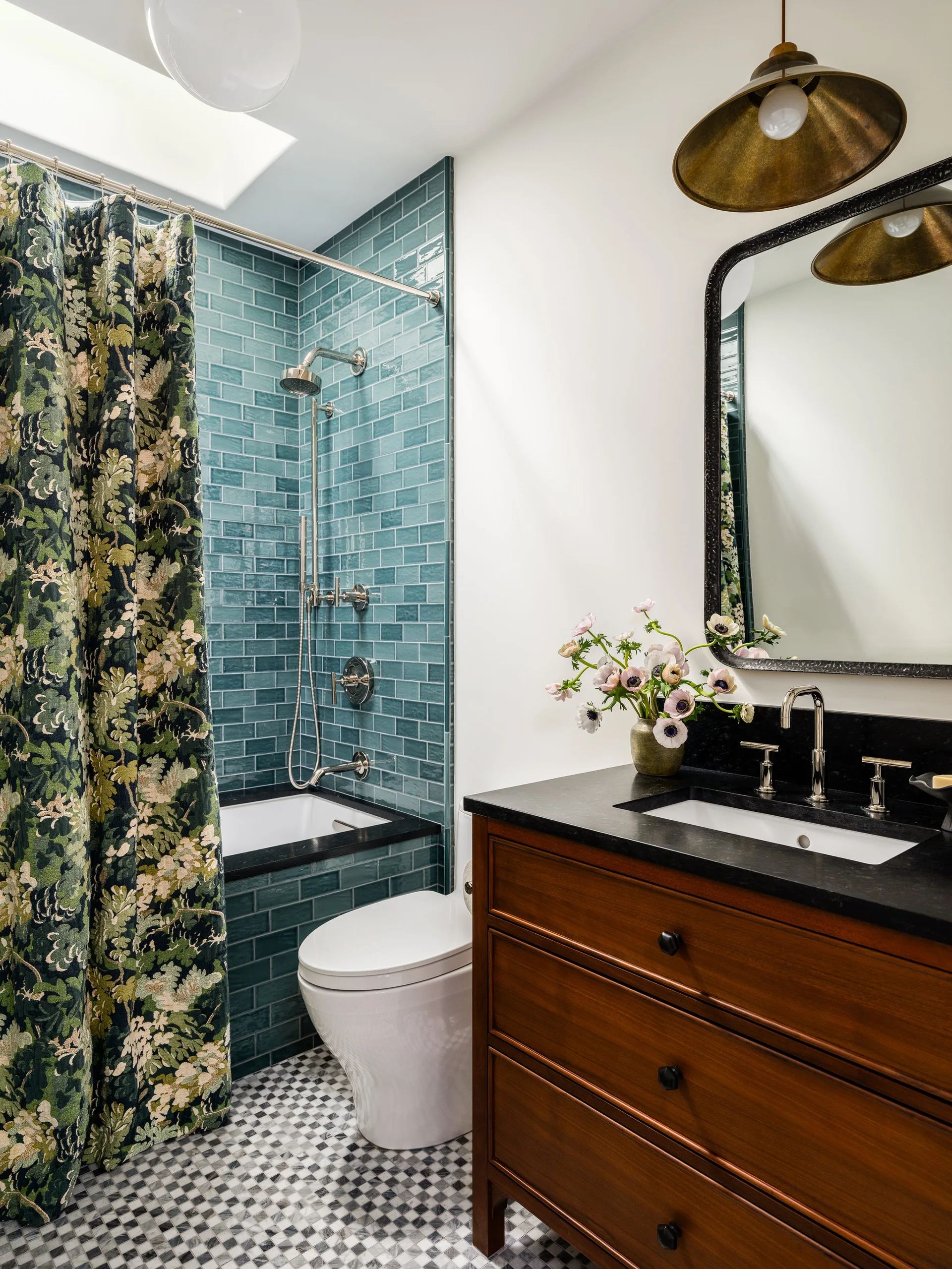 A bathroom featuring a warm wood vanity with a black countertop, a white toilet, and a shower lined with teal subway tile next to a floral curtain.