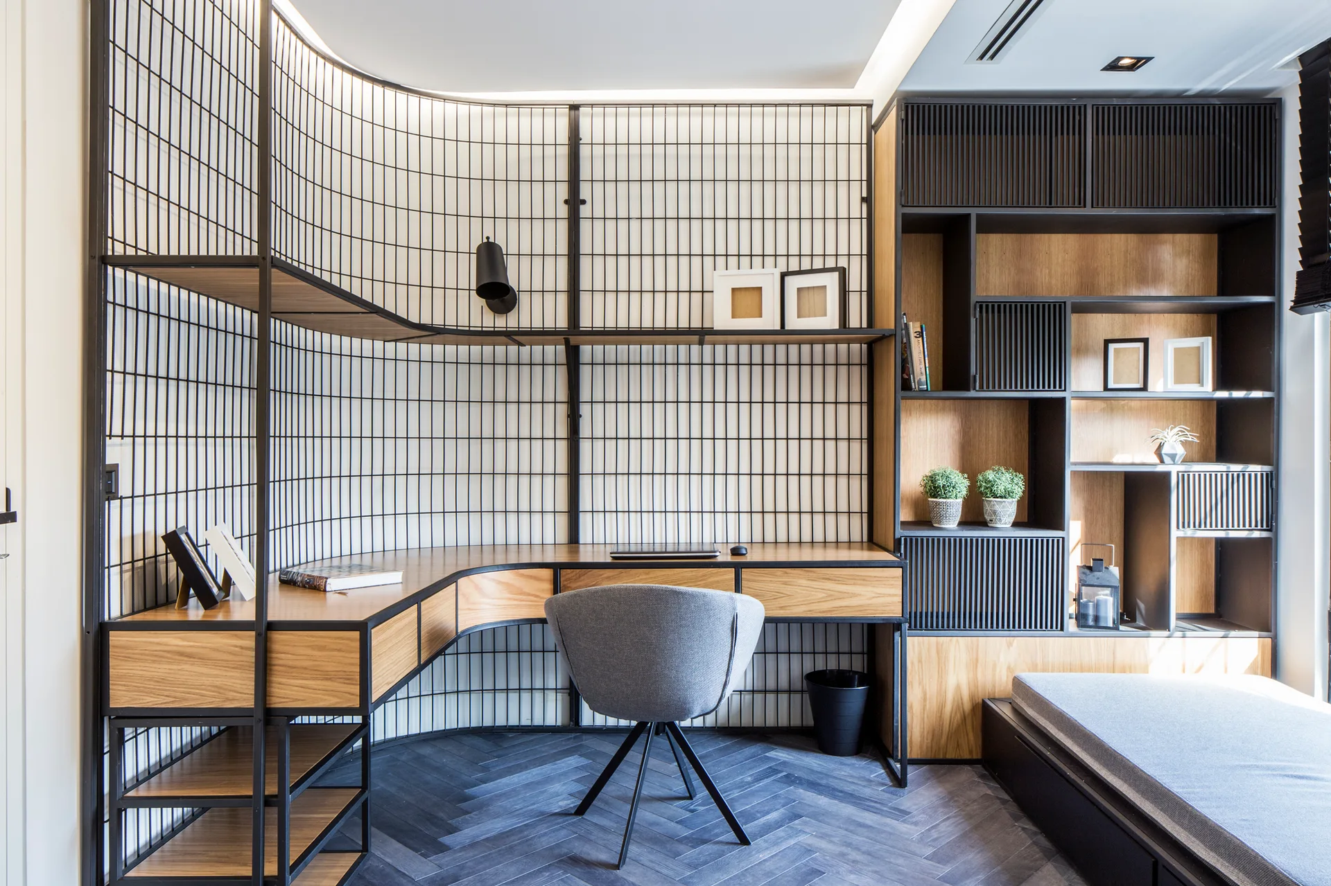 A modern home office features a curved wooden desk, a grey swivel chair, and a black wire grid screen partition wall.