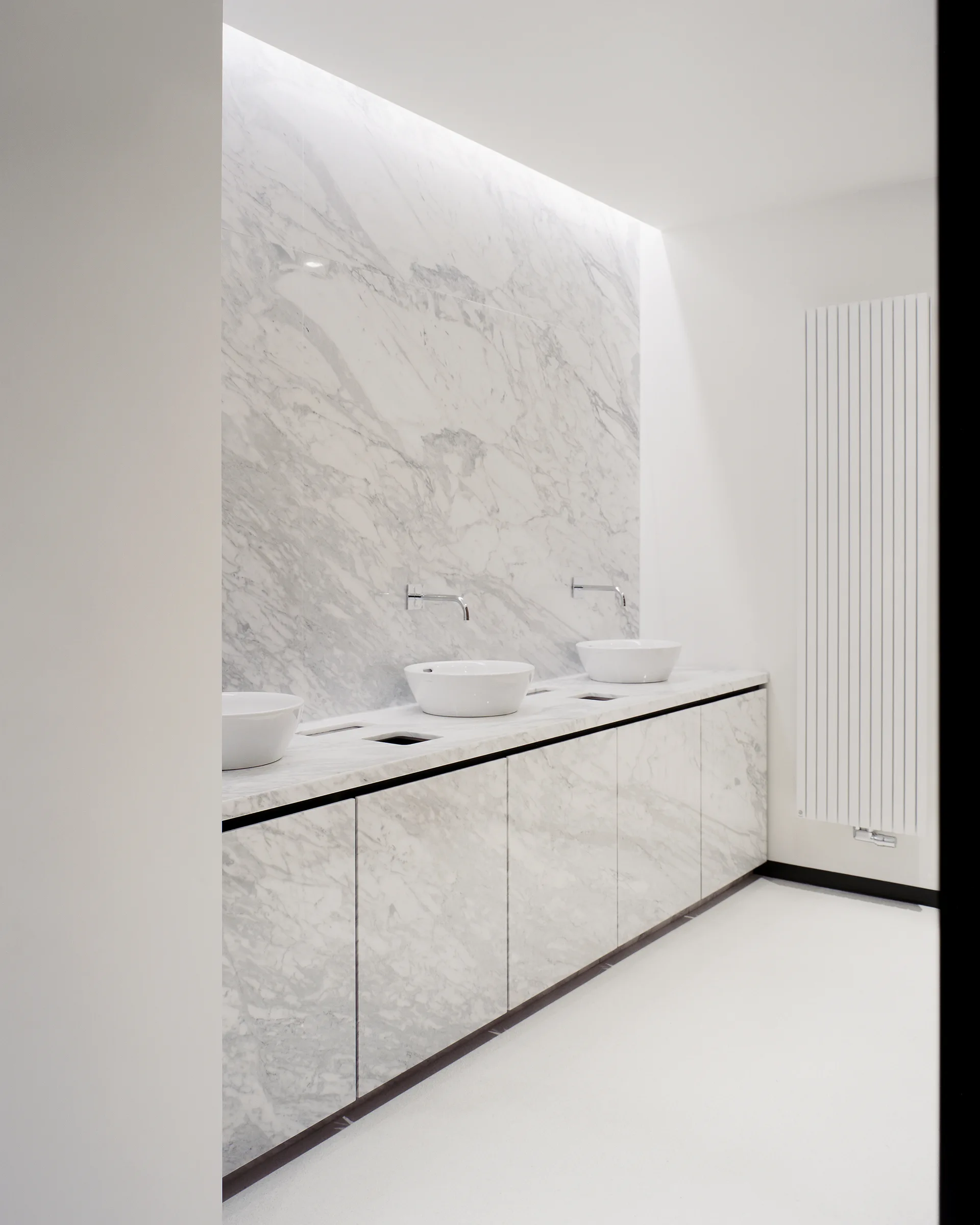 A minimalist bathroom vanity clad in white and grey veined marble features three vessel sinks under wall-mounted chrome faucets.