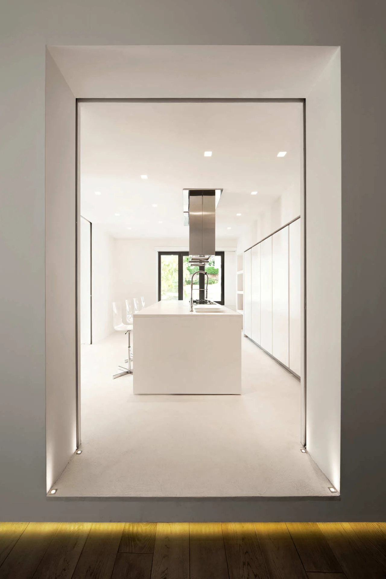 A minimalist white kitchen viewed through a rectangular doorway, featuring a central island, full-height seamless cabinetry, and floor-level uplighting.