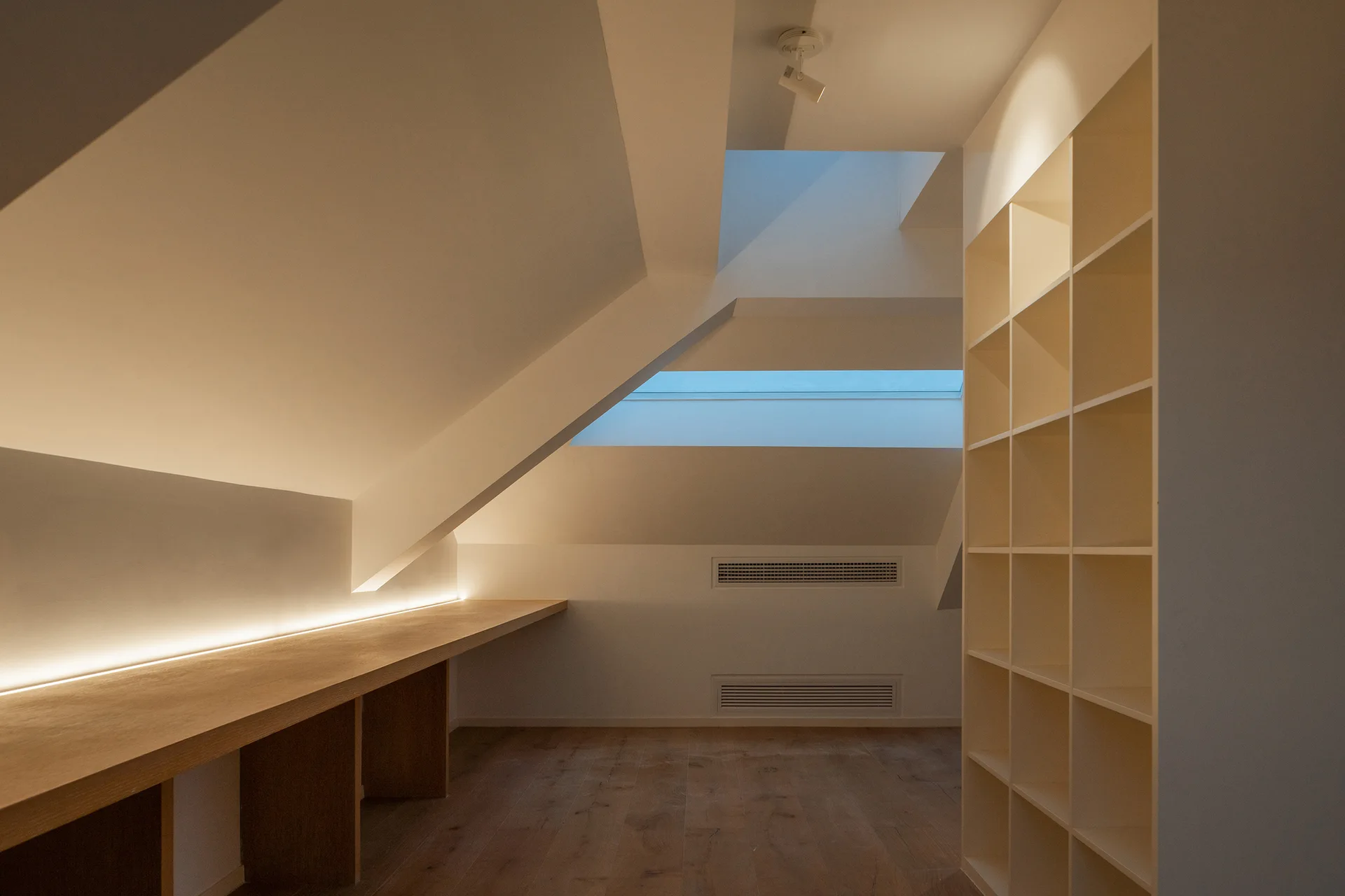 A minimalist attic room features a long built-in wooden desk with illuminated LED backlighting under a sloped ceiling and a white grid shelving unit.