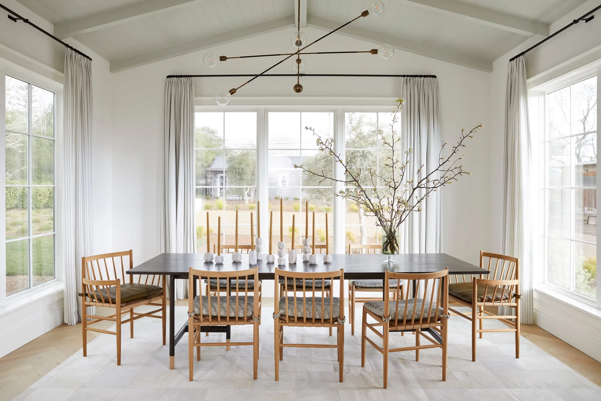 A light-filled dining room features a long dark table, light wood spindle-back chairs, a modern Sputnik chandelier, and large windows with cream drapes.