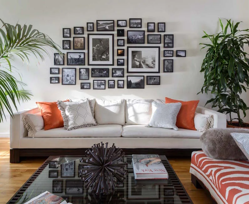A neutral living room features an off-white sofa with orange pillows beneath a large gallery wall of black-framed photographs.