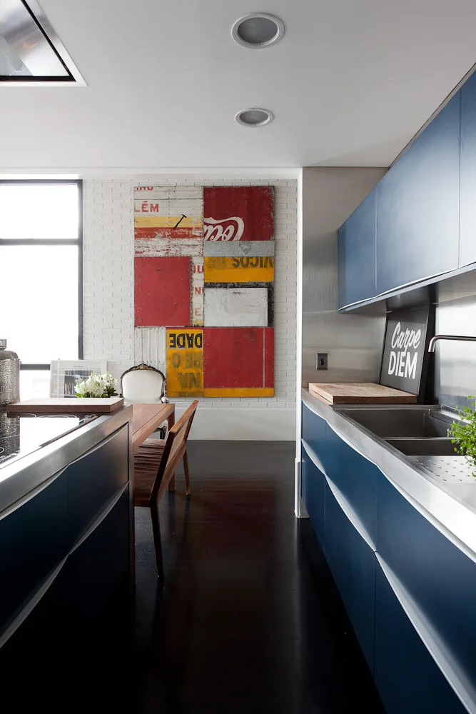 A modern kitchen features matte blue cabinets, stainless steel countertops, dark wood flooring, and a dining table next to a white brick wall.